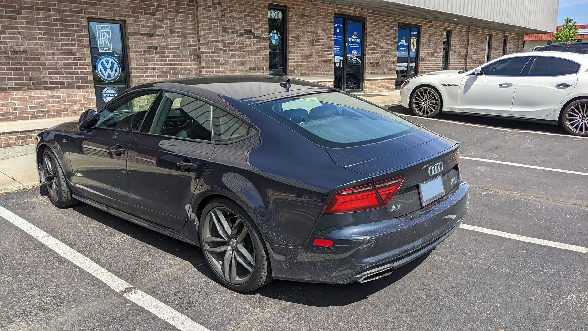 Dark gray Audi A7 parked in front of a brick building; another white car is parked beside it.