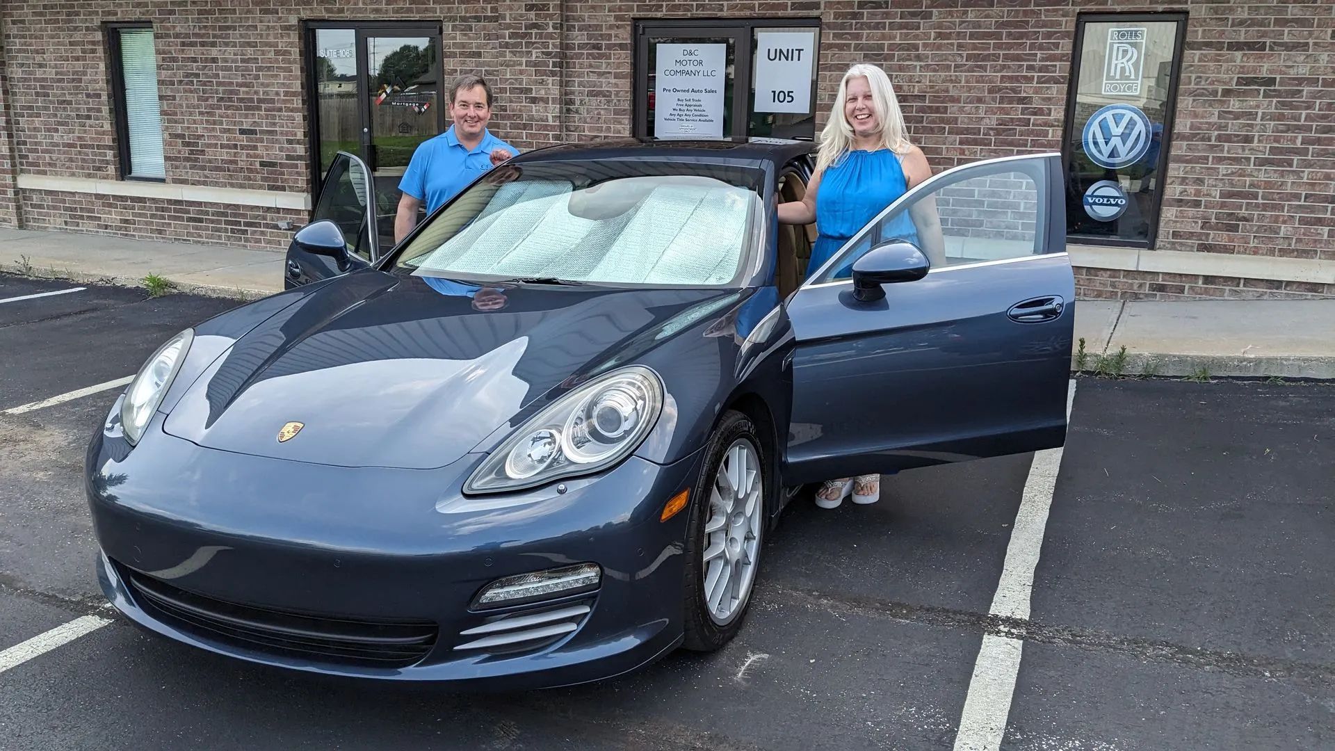 Man and woman stand by a blue Porsche with open doors in front of a building.