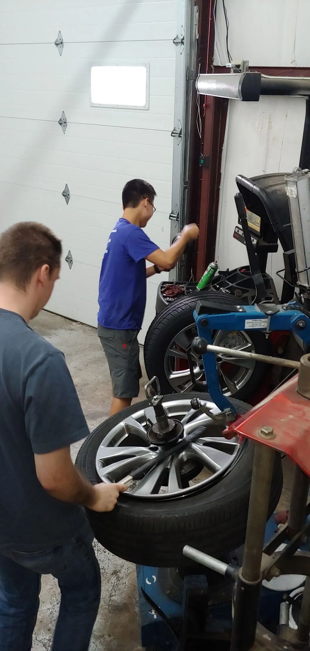 Two people changing a tire in a garage. One holds the tire, and the other works on a machine.