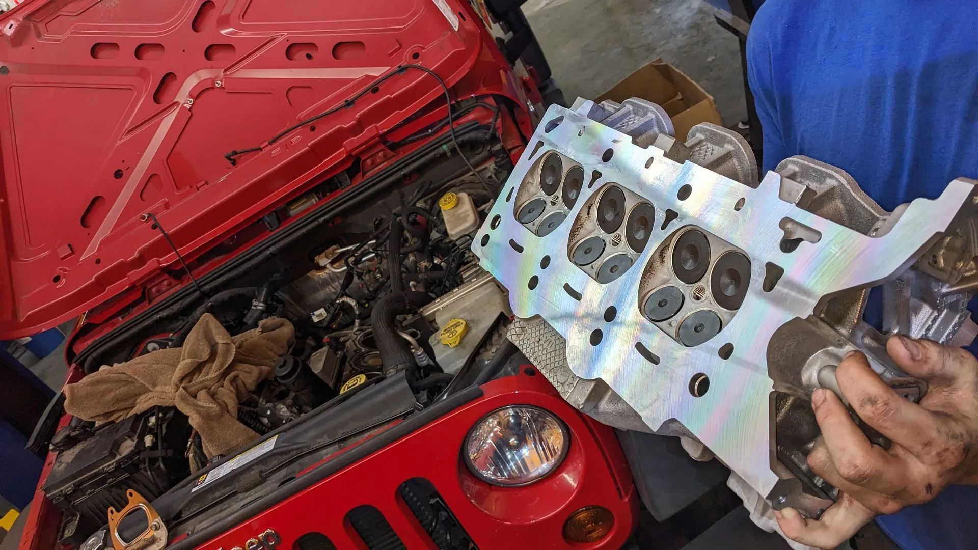 Mechanic holding a cylinder head next to a red Jeep with the hood open, engine visible.