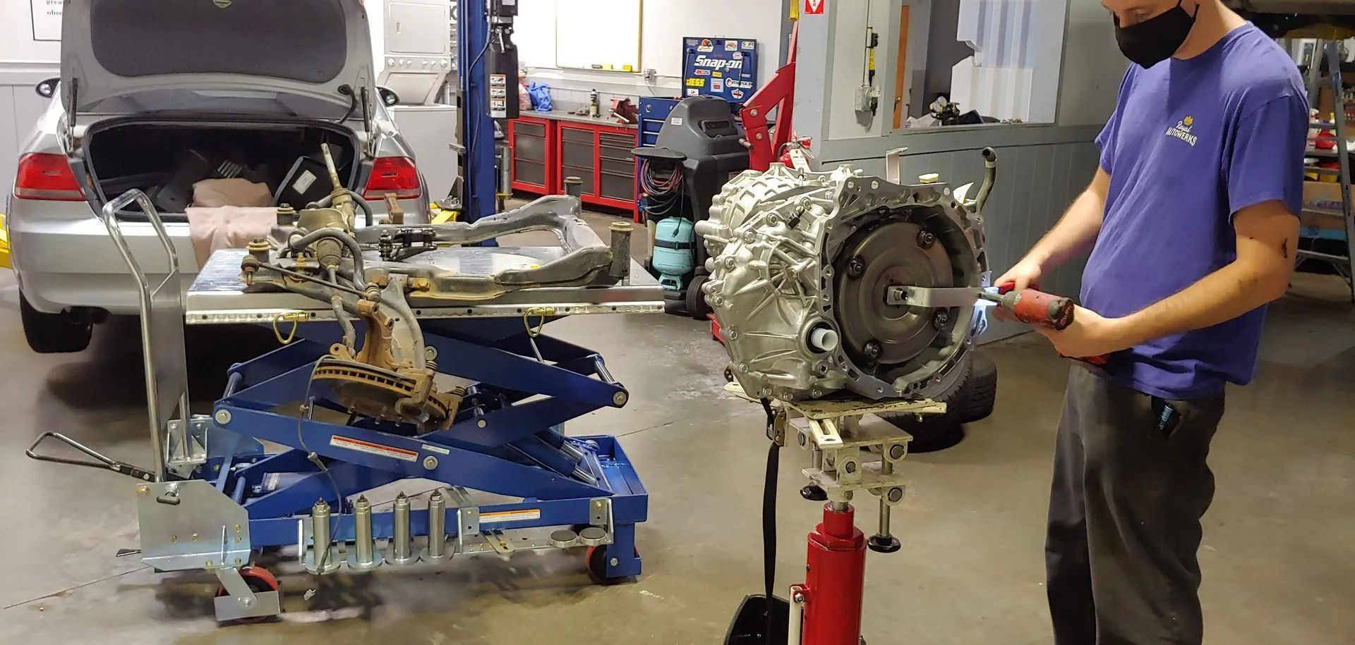 Mechanic working on a transmission in an auto shop. A car is visible with open trunk.