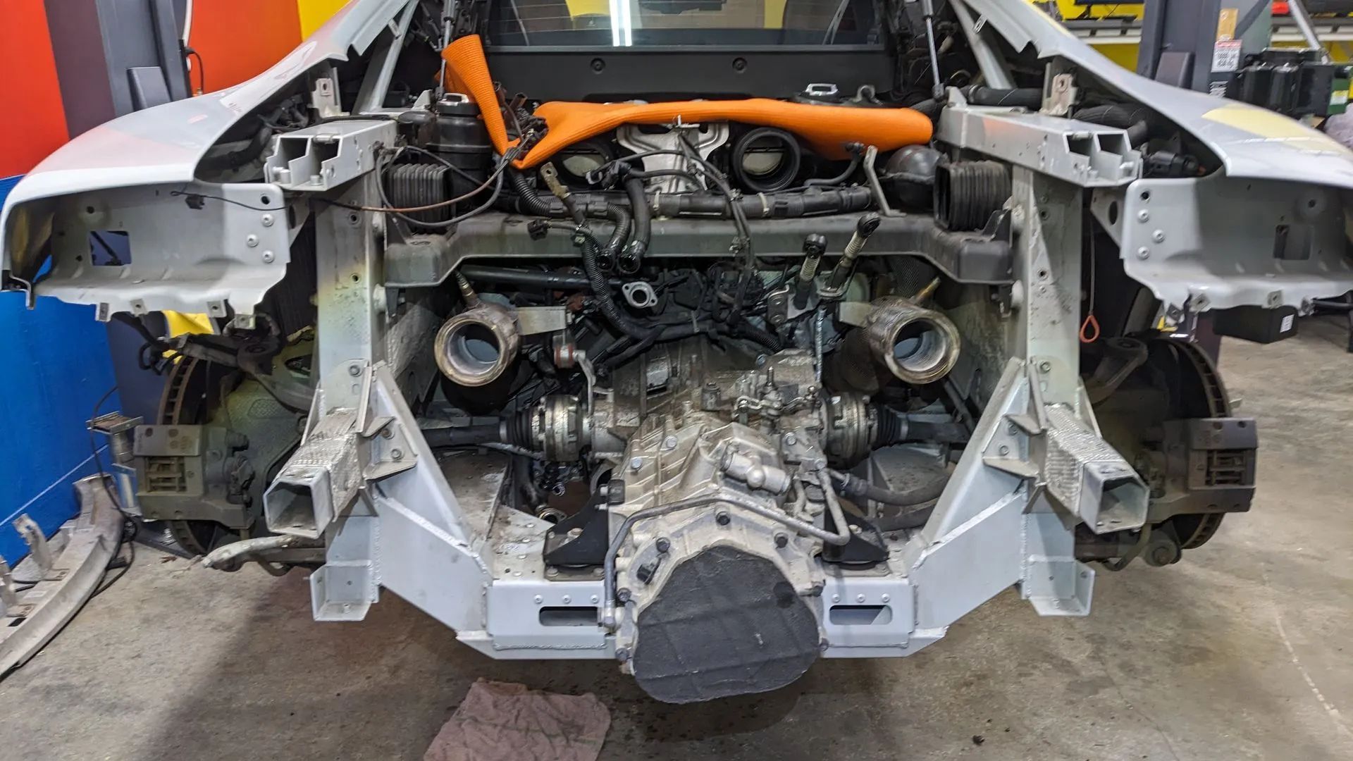 Car's front end disassembled, engine exposed. Silver frame, black and orange components, wheels visible. Workshop setting.