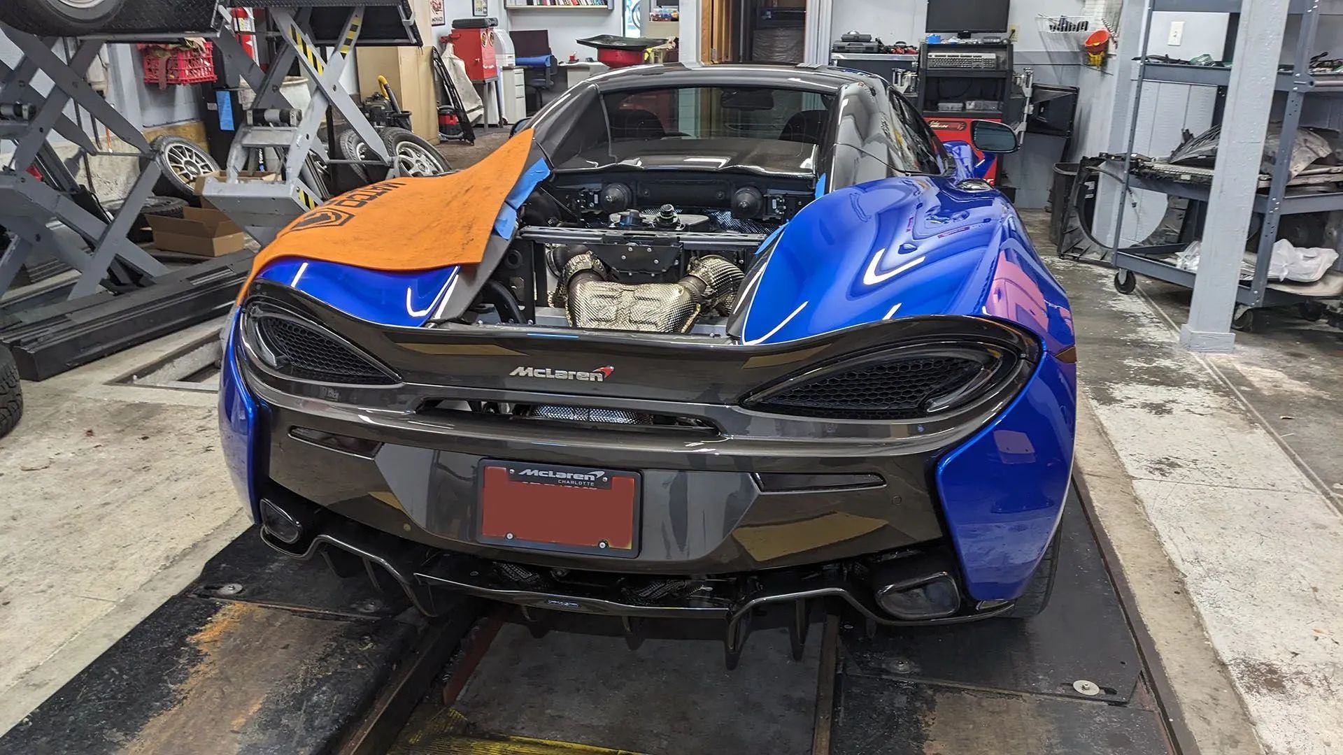 Blue McLaren sports car in a garage, engine bay open, covered in orange and blue masking.