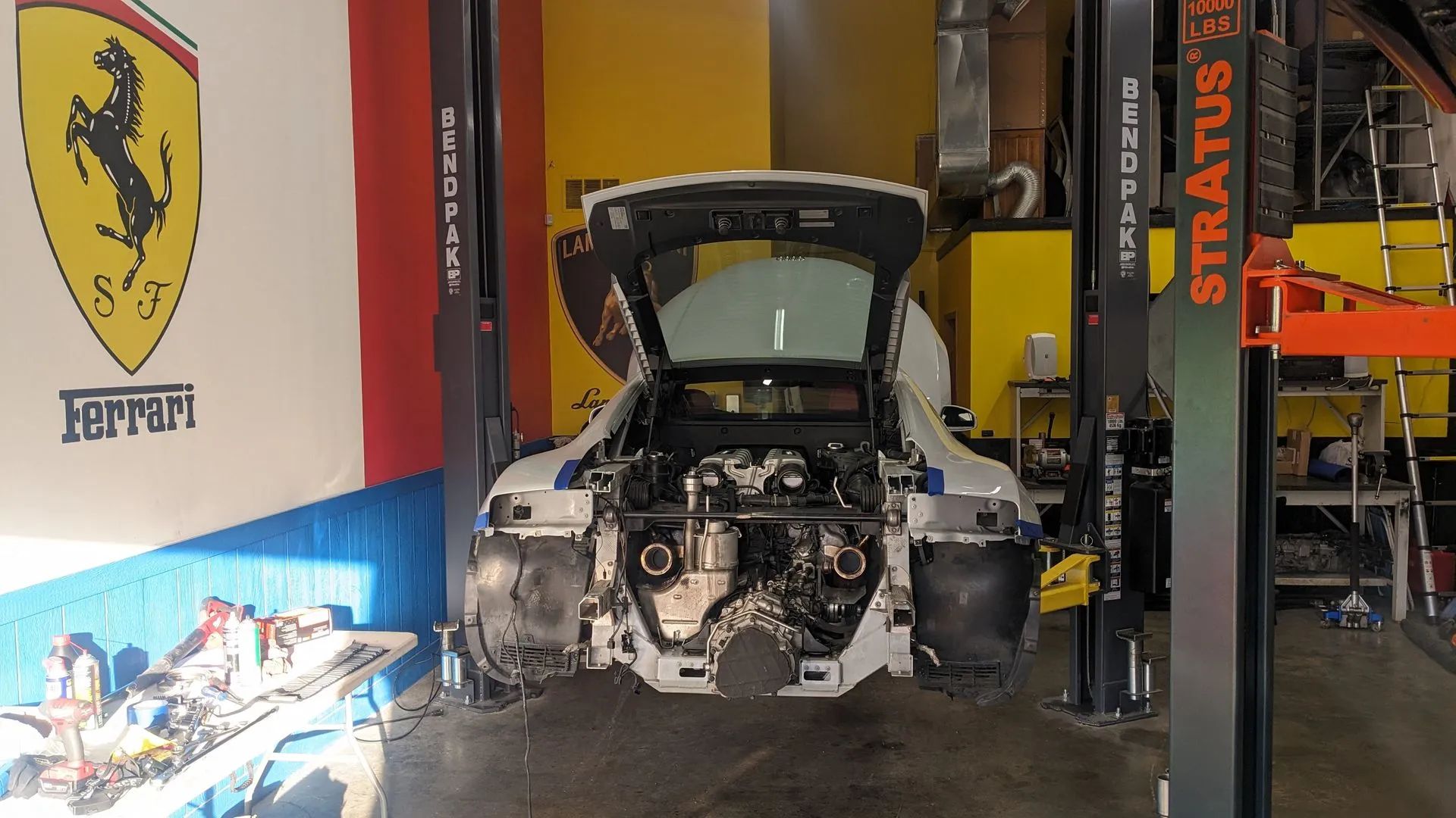 White sports car undergoing repair in a garage, with the hood and doors open. Ferrari logo on the wall.