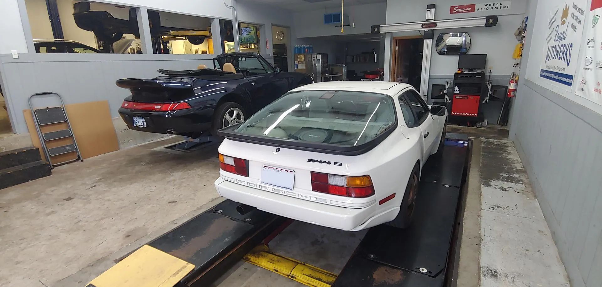 White and black cars in a garage. One on a lift, one on the floor.