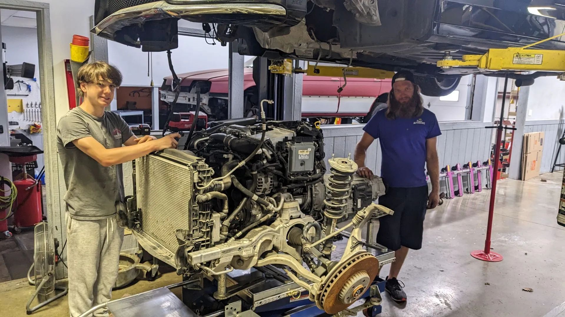 Two people stand near a car's engine, possibly in a repair shop. One touches the engine, the other stands.