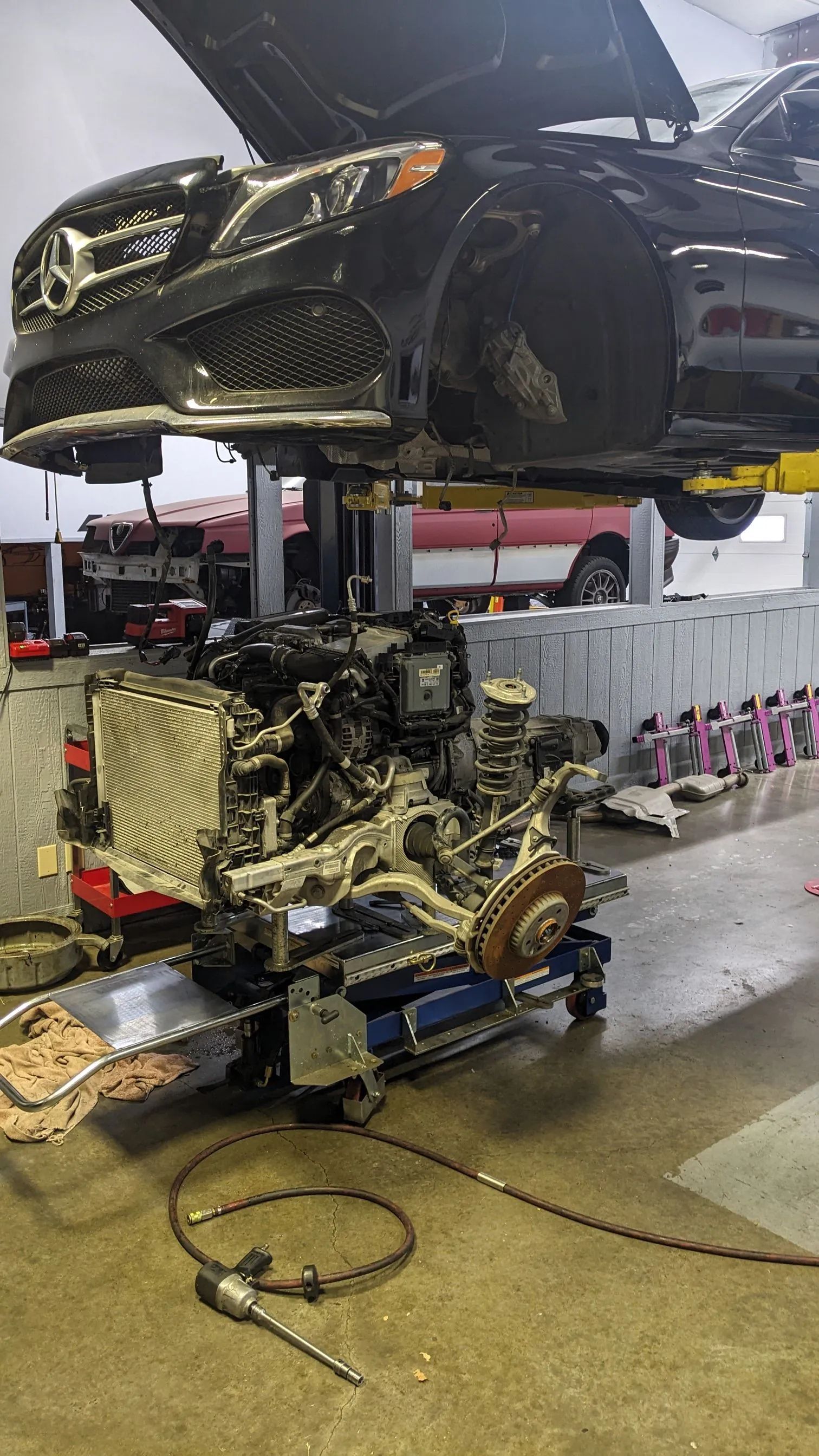 Car's engine removed, suspended on a lift in a repair shop. Black car above; engine on a stand below.