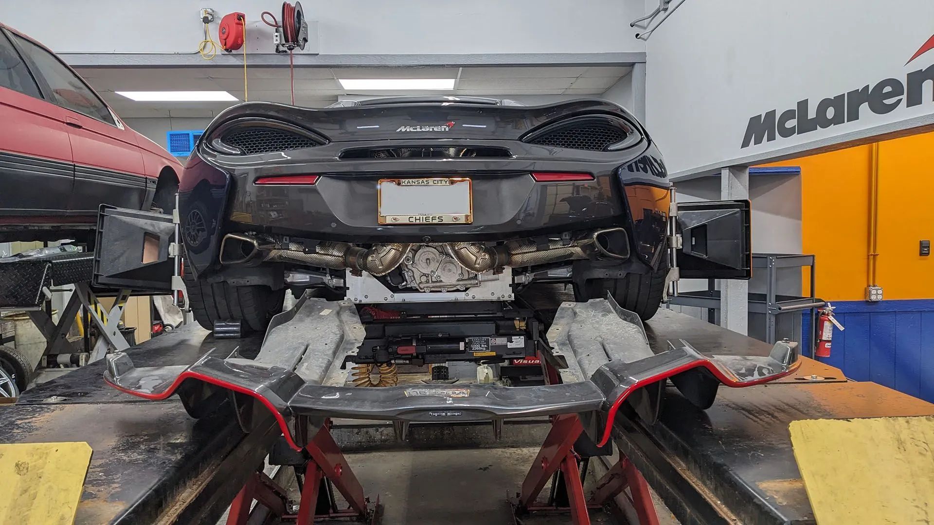 A black McLaren sports car is elevated on a lift in a garage. Exhaust visible, with a red car next to it.