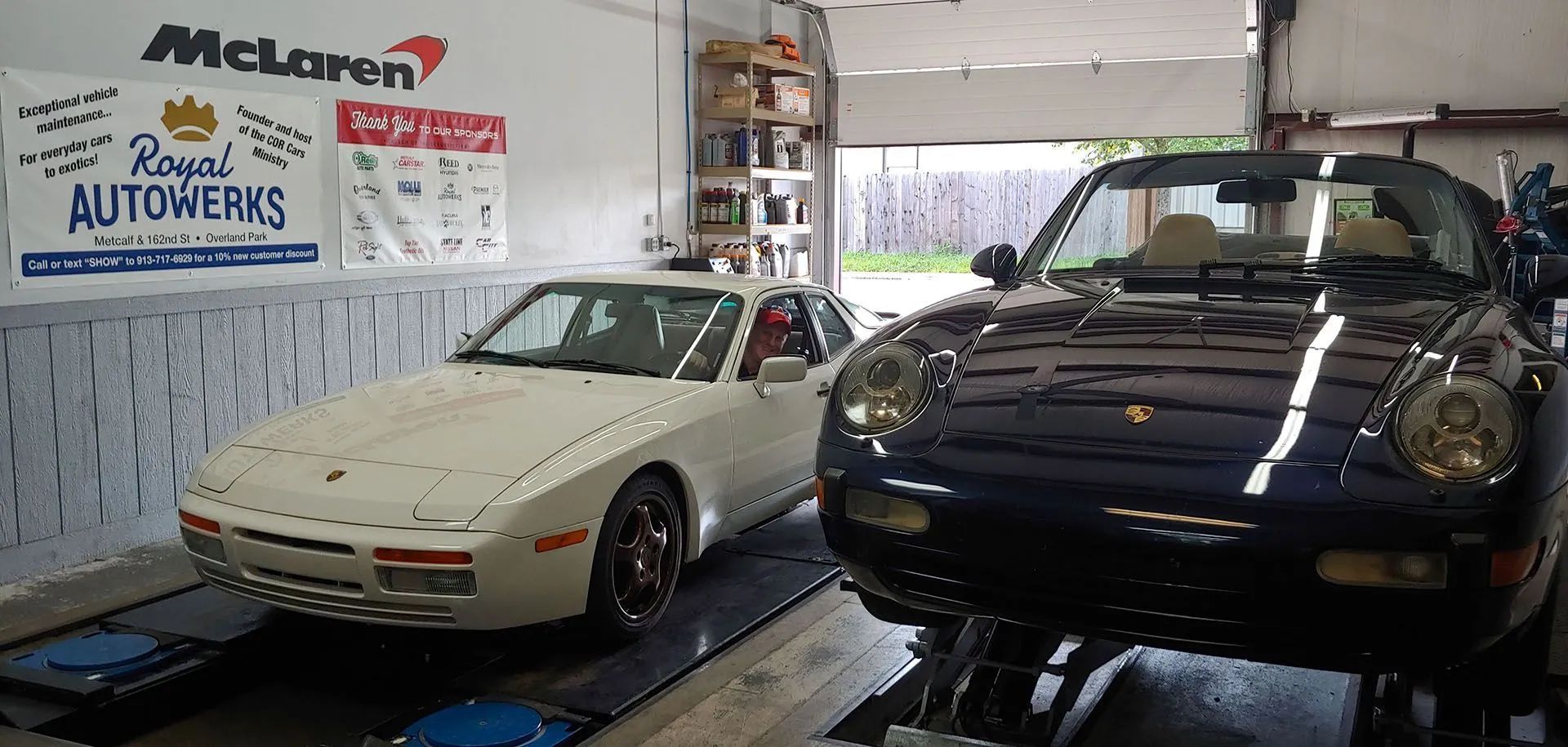 Two Porsches in a repair shop. One white, one blue convertible on a lift. Shop name visible on the wall.