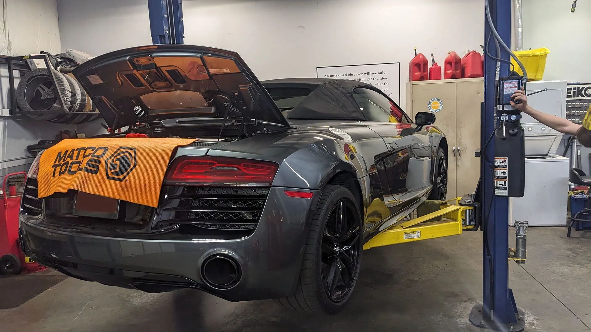 Gray Audi R8 on a yellow car lift, hood open. A person operates the lift in a garage.