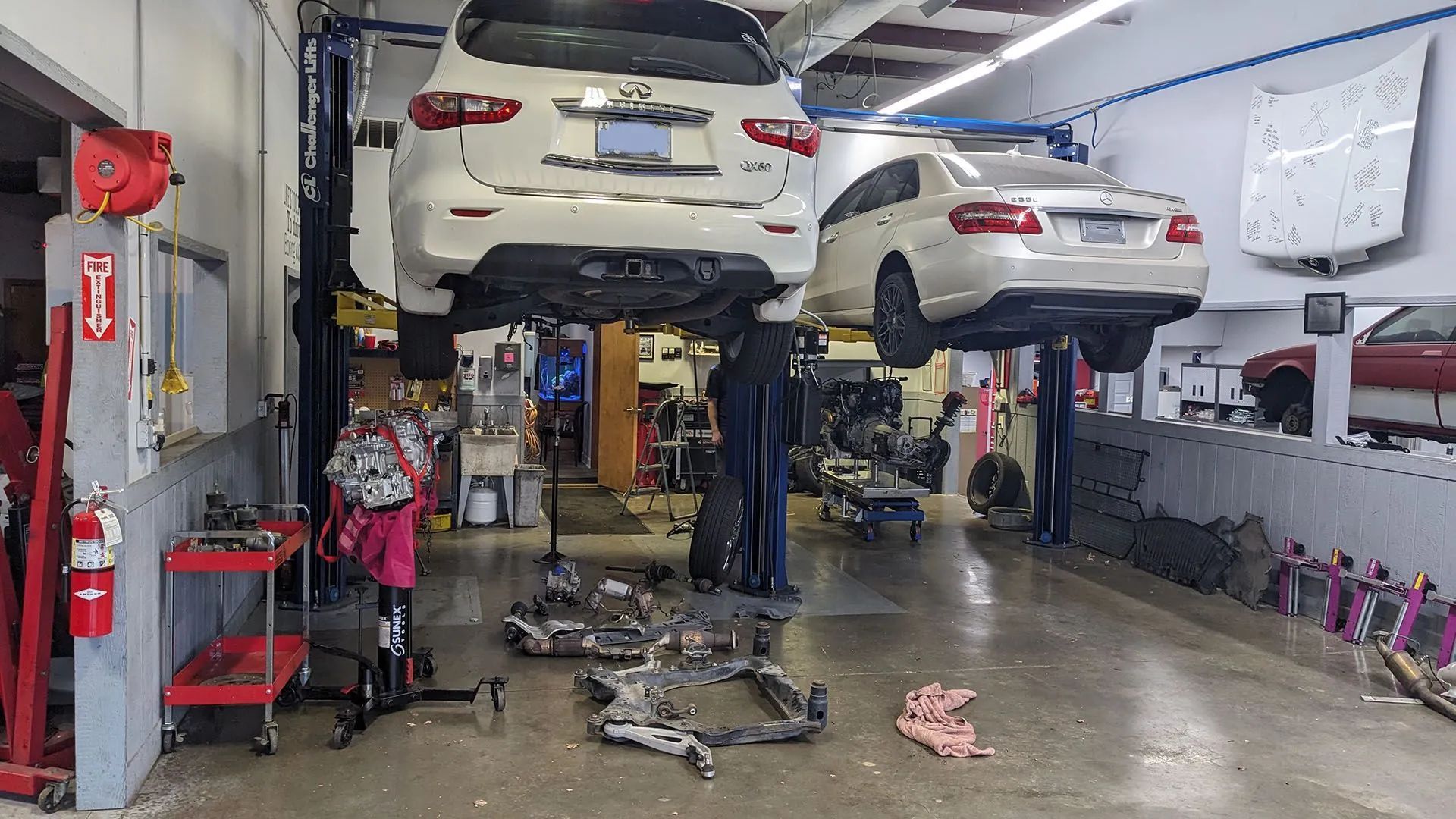 Cars on lifts in a repair shop. Parts are scattered on the floor. People are working on the vehicles.