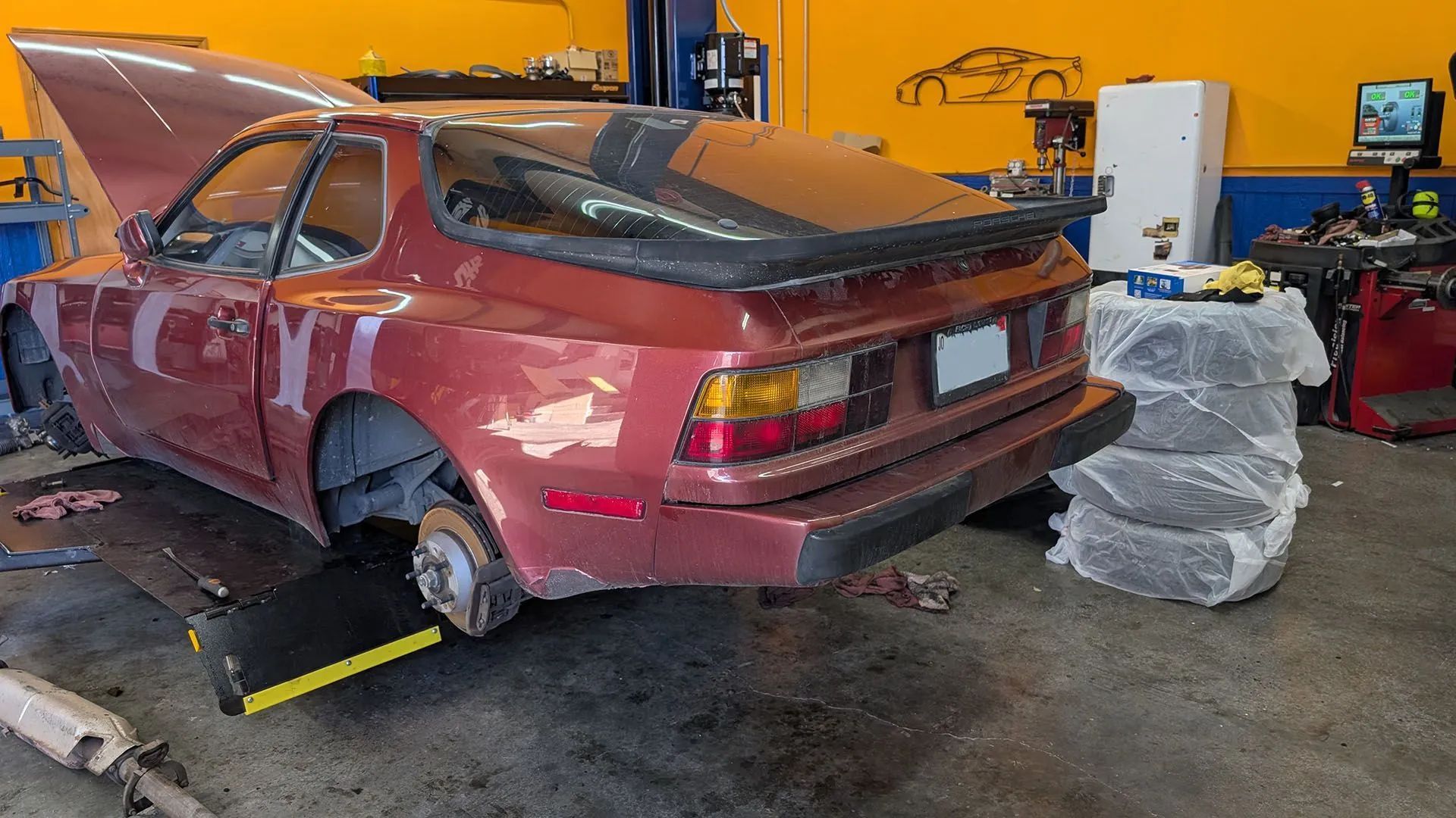 A maroon Porsche 944 being worked on in an auto repair shop. Hood up, rear tire removed.