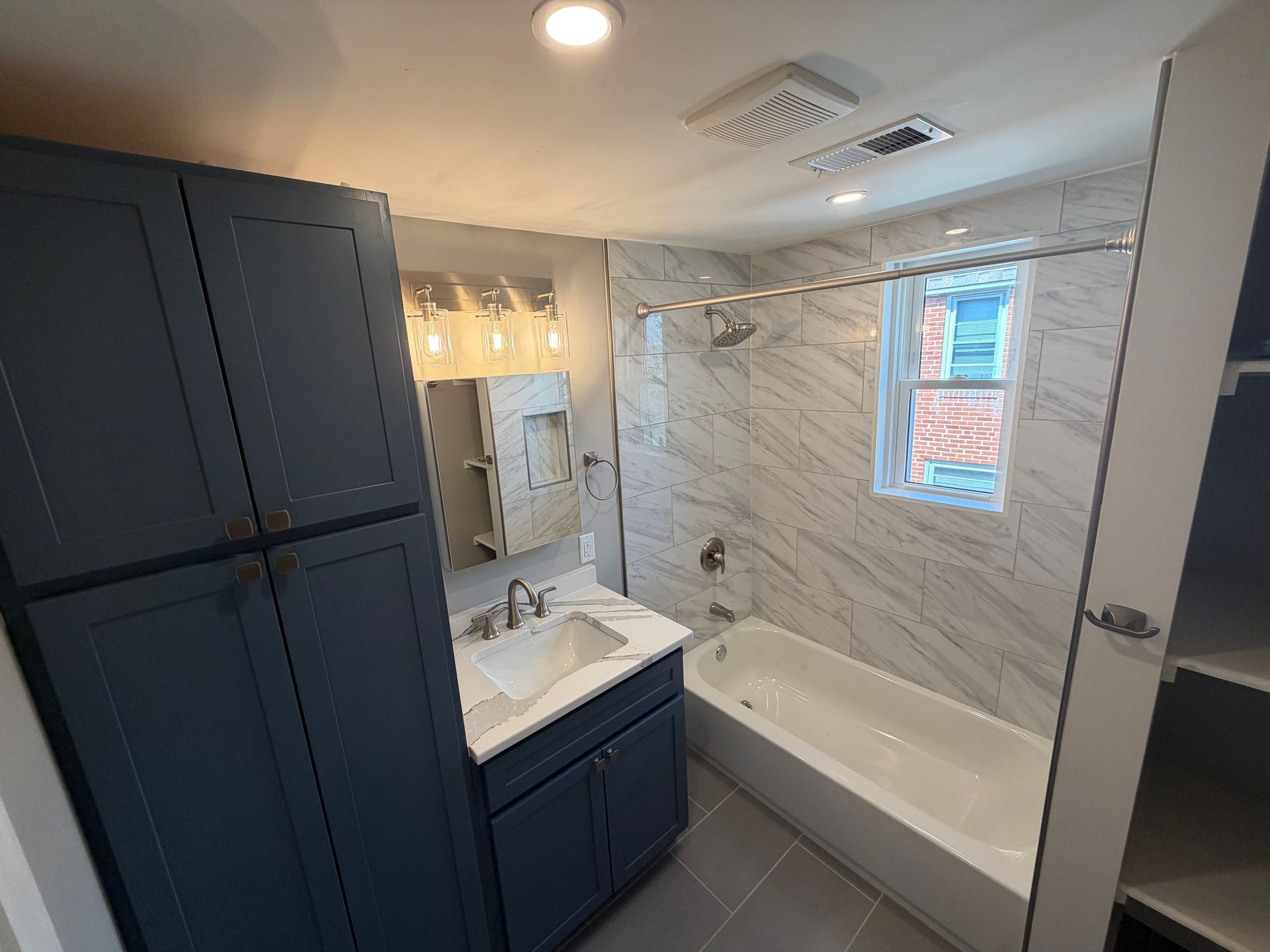 Modern bathroom with dark vanity, marble tile, bathtub, mirror, and window lighting