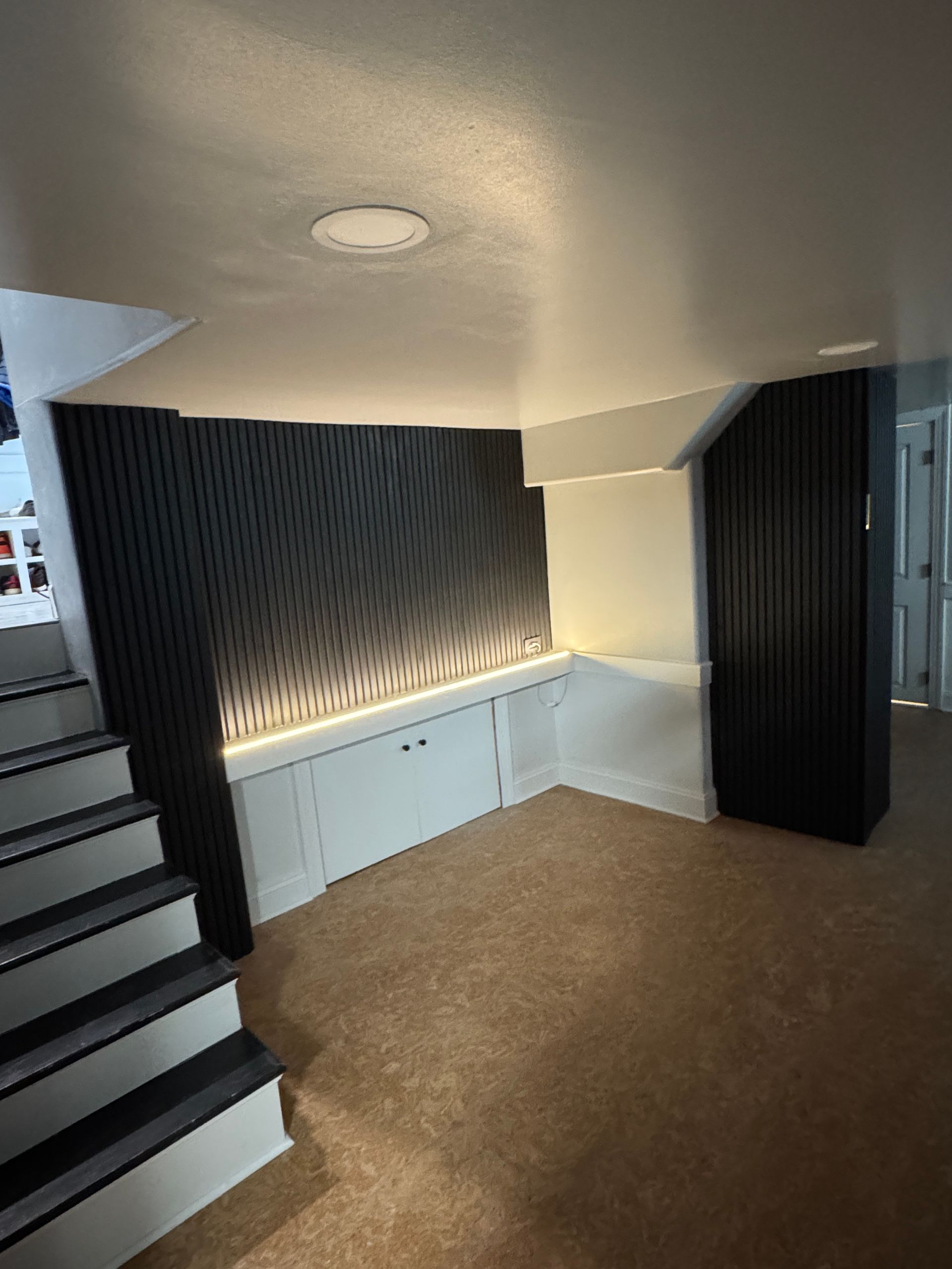Basement with stairs, carpet, and built-in white cabinets. A dark gray paneled wall is center.
