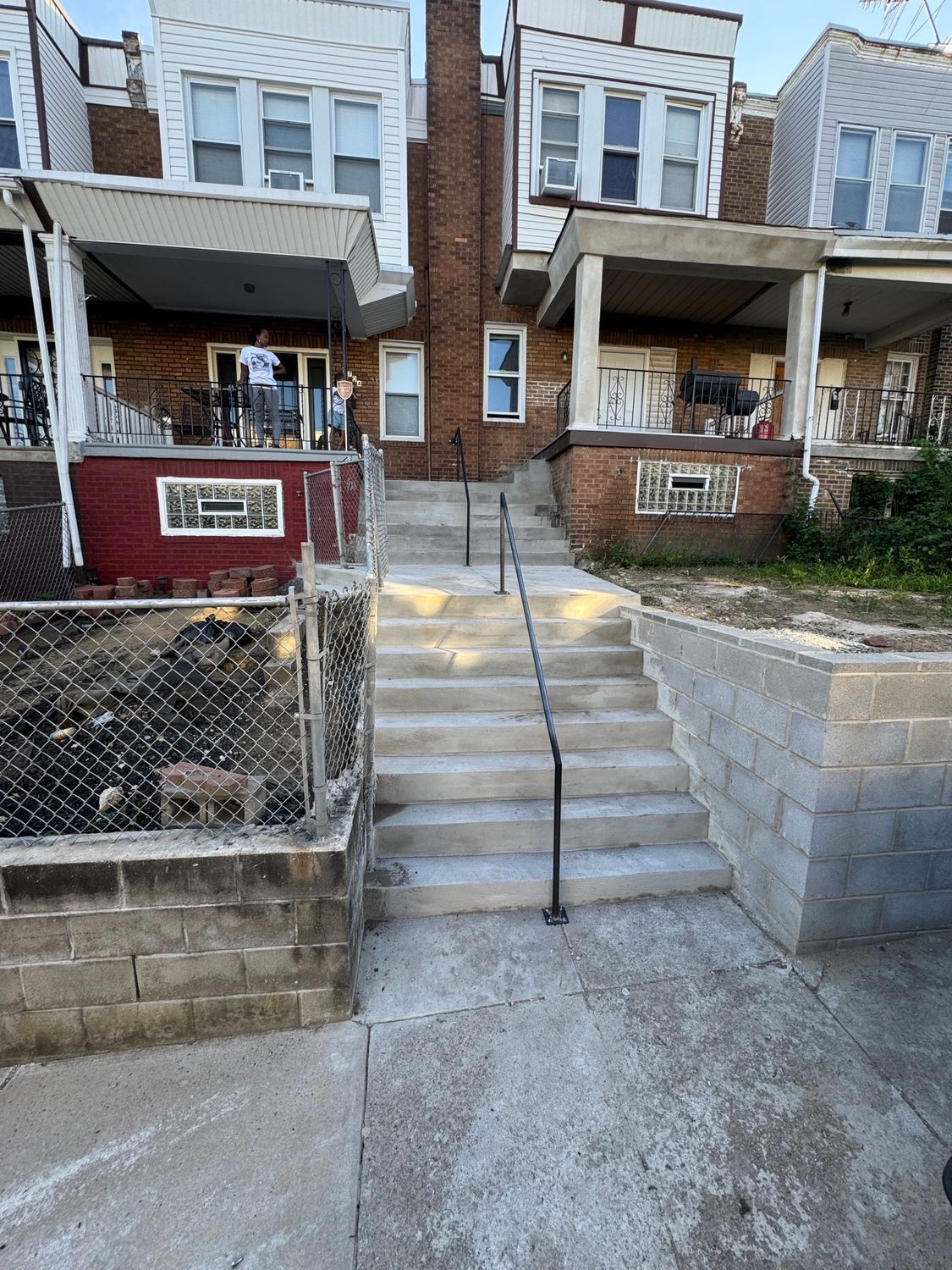 Concrete steps lead to row houses. Black handrail. Brick and concrete walls. People on porches.