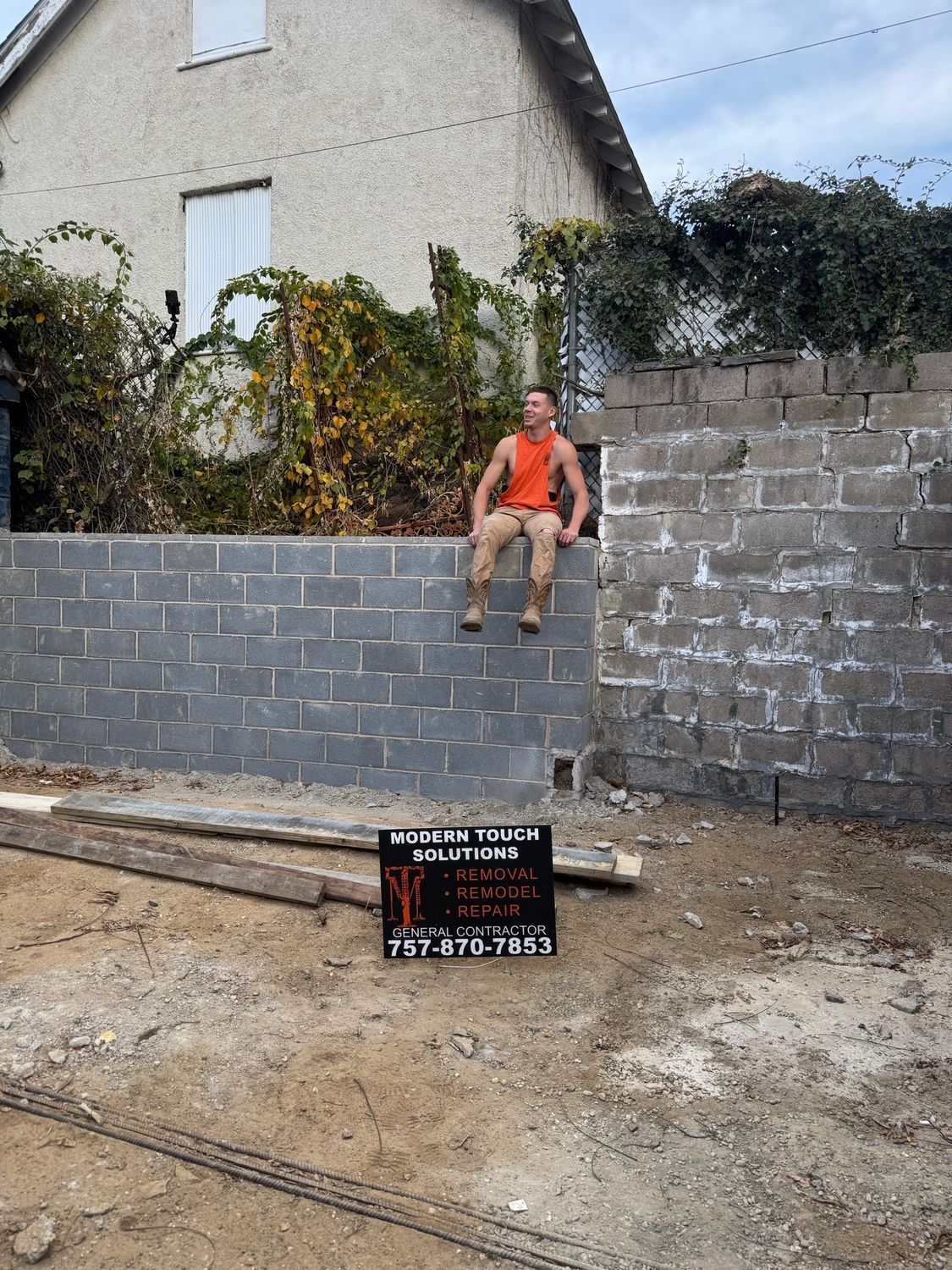 Man sits on a gray cinder block wall under construction. Sign in front. House in background. Cloudy sky.