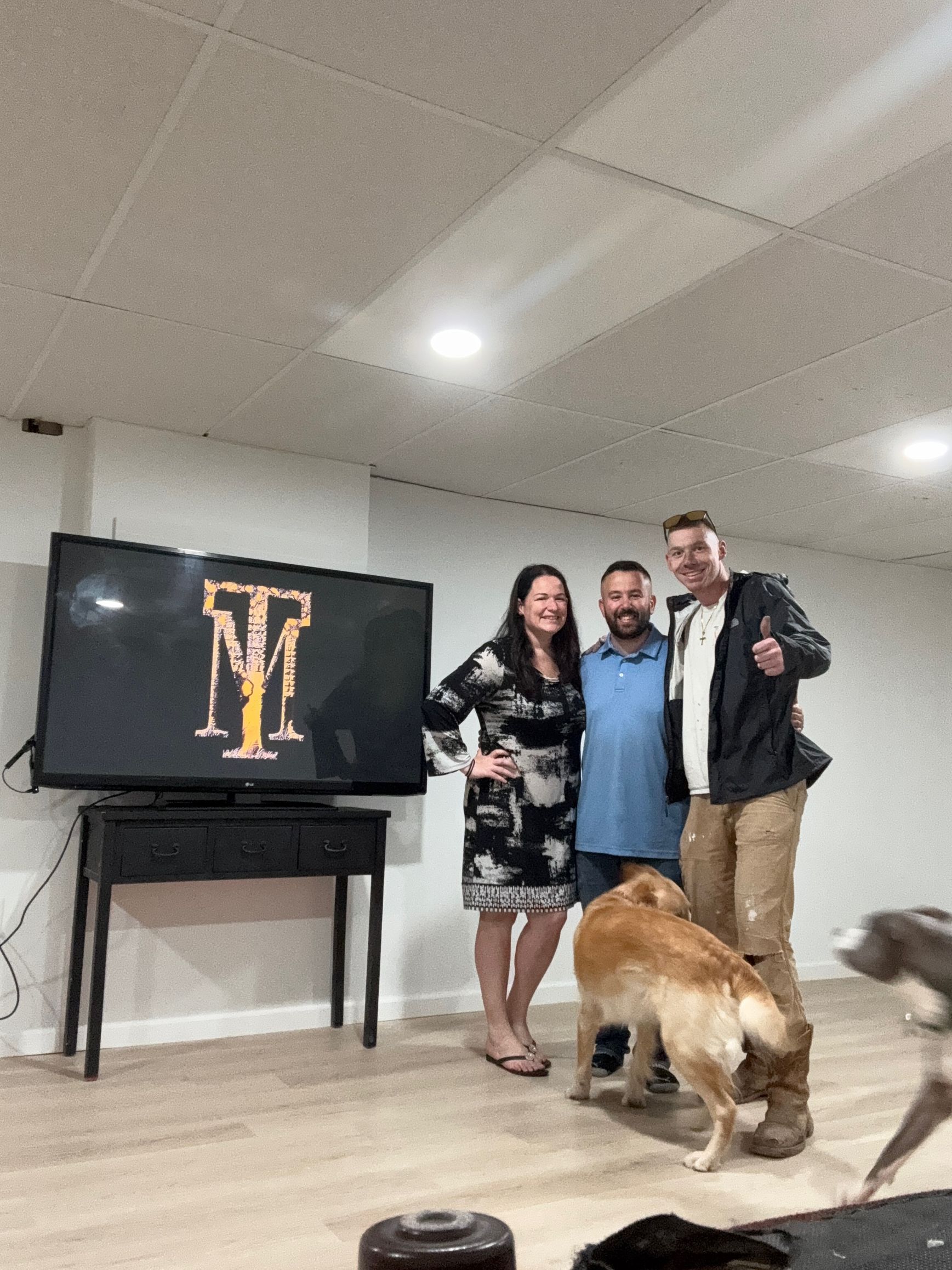 A group of three people and two dogs pose in front of a TV with a logo.
