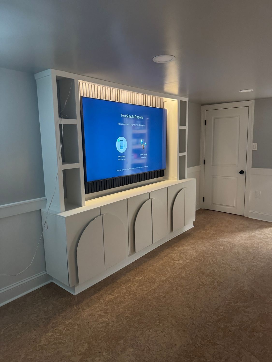White built-in media center with TV. Includes cabinets, shelves, and lighting. Room with white walls and brown carpet.