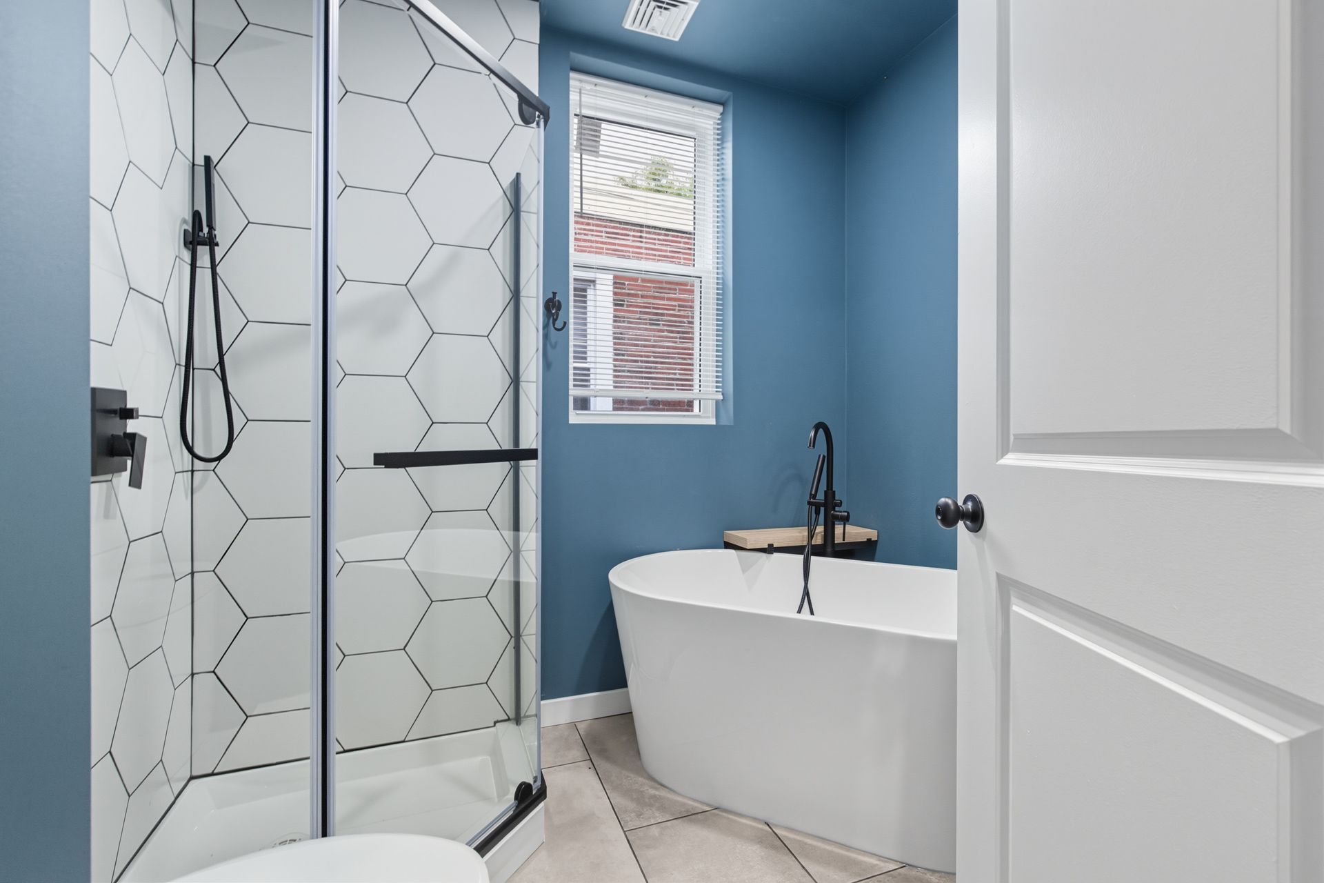 Bathroom with blue walls, white bathtub, hexagon tile shower, and a window with a view of brick buildings.