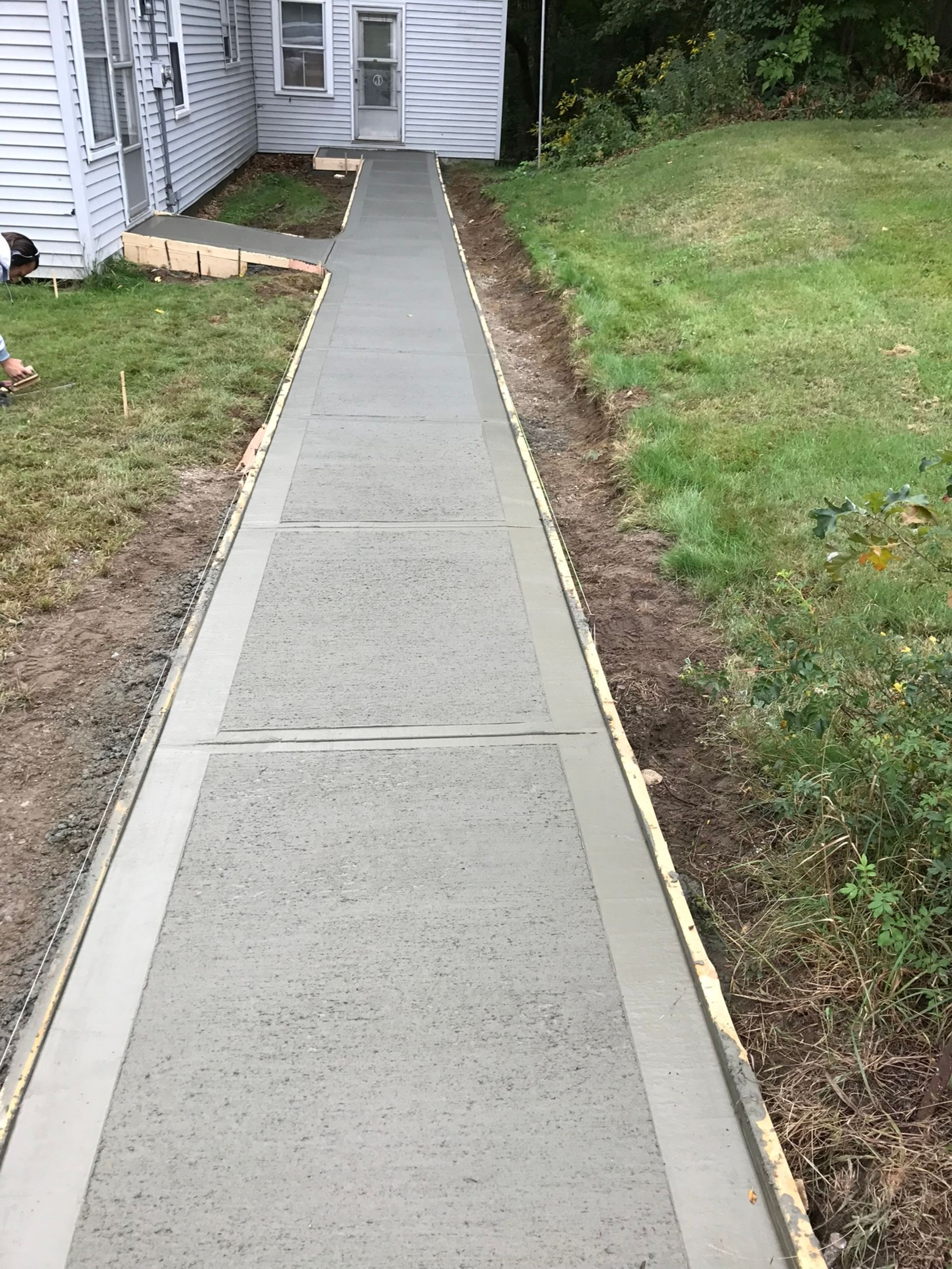 A newly poured concrete walkway leads to the side entrance of a house, framed by grass and wooden forms.