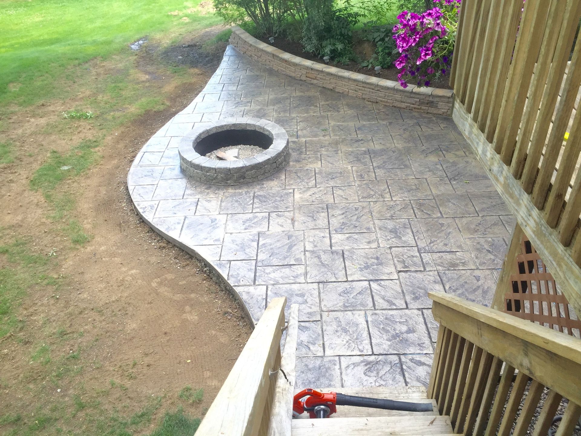 A stone-patterned stamped concrete patio with a circular fire pit, viewed from the wooden steps of an elevated deck.