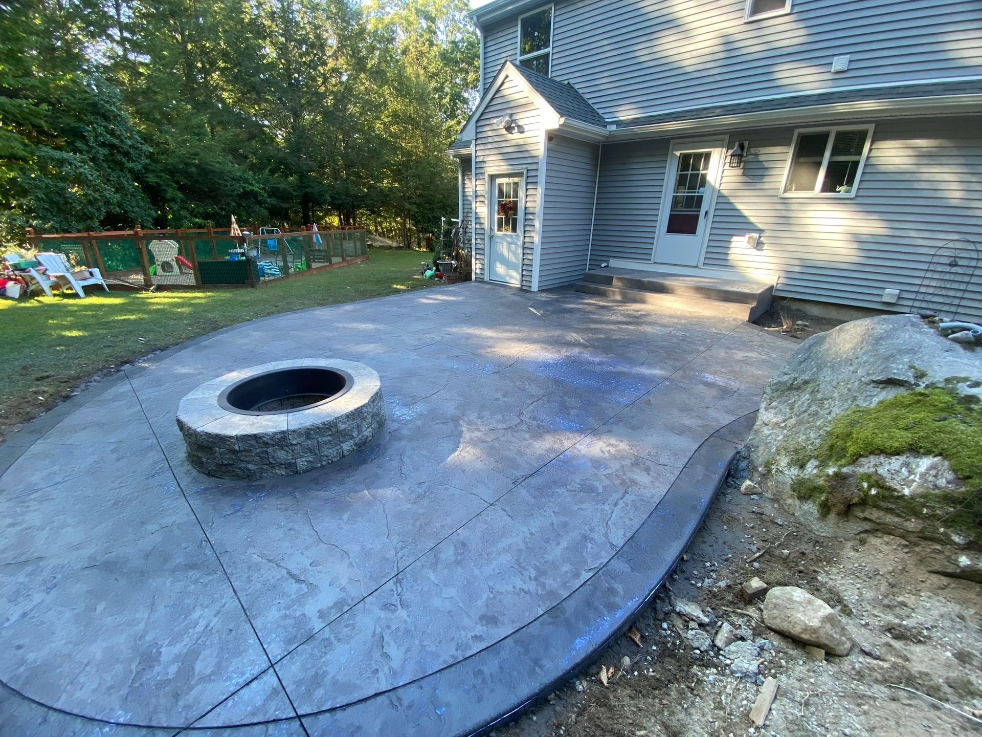 A grey, stamped concrete patio with a circular stone fire pit sits in a backyard next to a grey house.