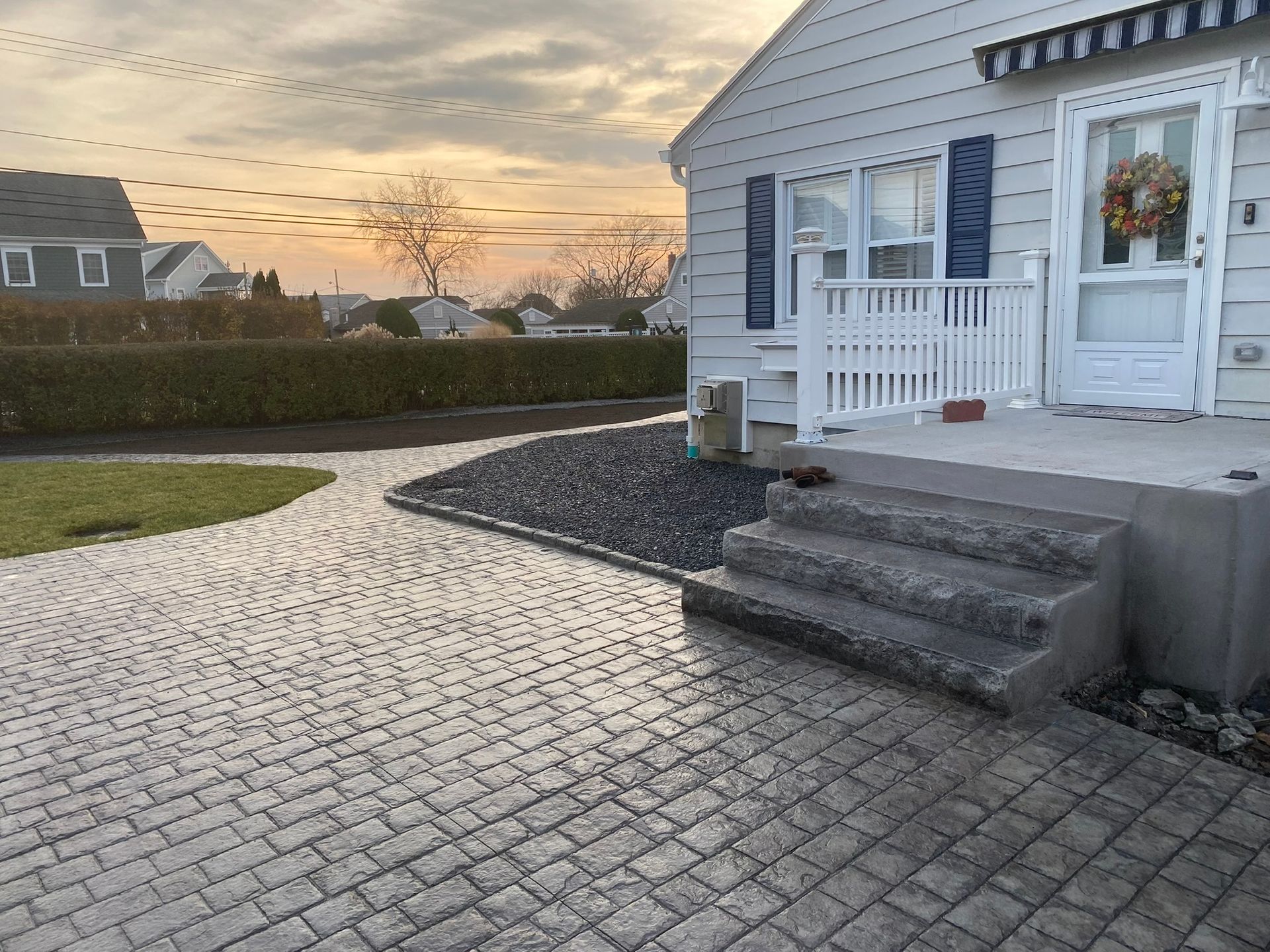 A paved walkway and patio lead to the front steps of a white house with blue shutters at sunset.