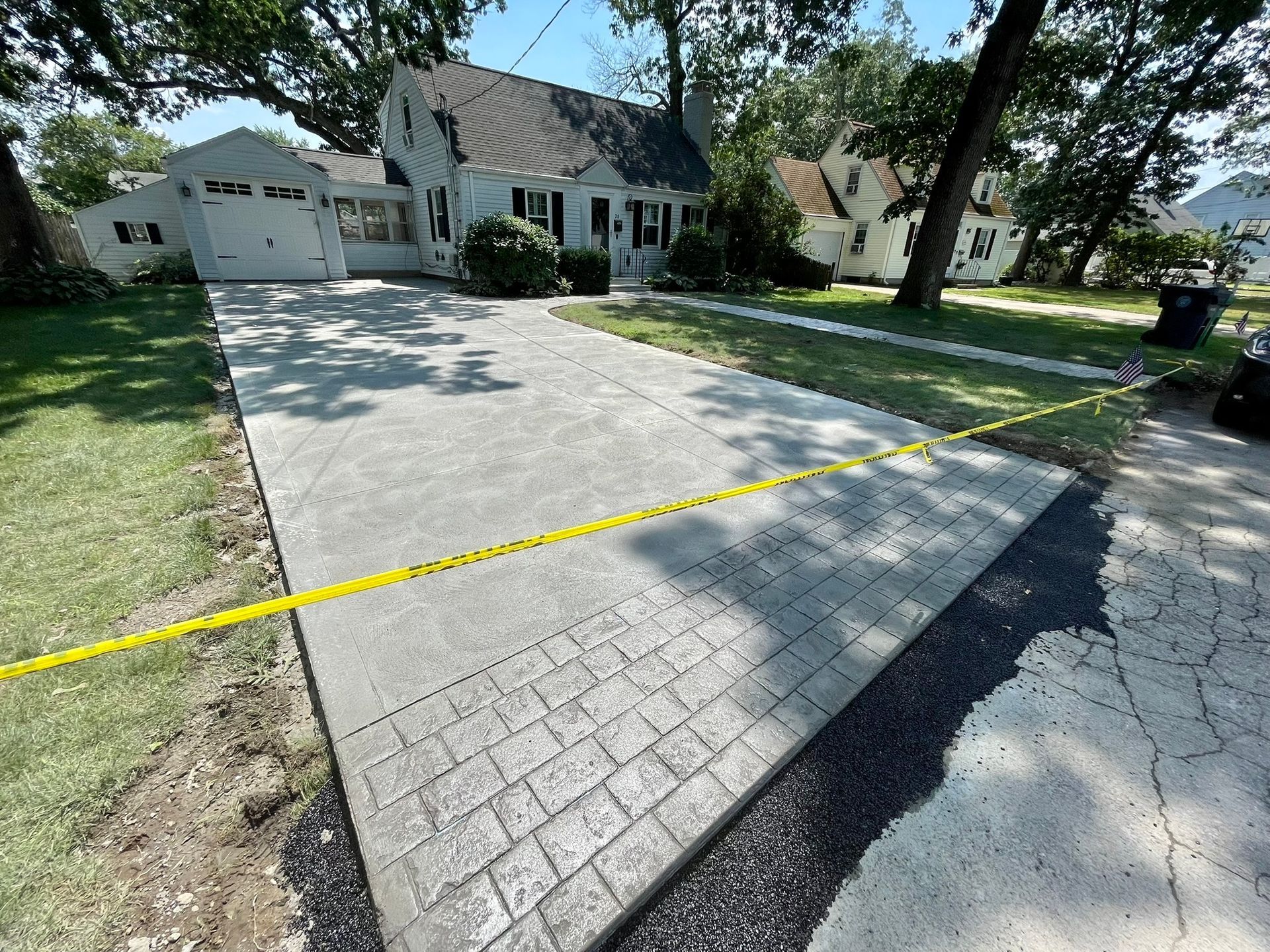 A paved driveway with a patterned border leads to a suburban house with a garage, blocked off by yellow caution tape.