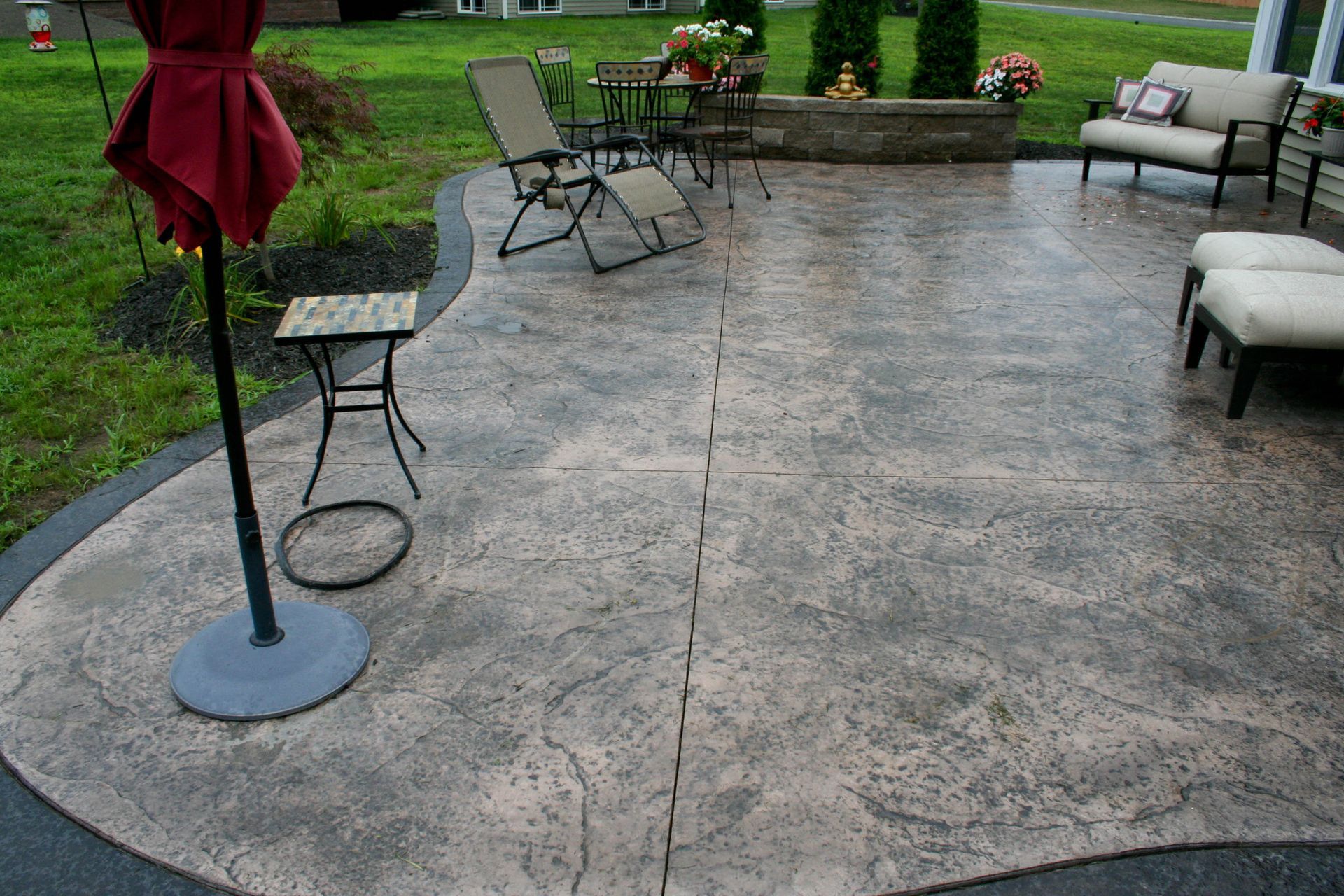 A stamped concrete patio featuring a set of patio furniture, including a lounge chair, a sofa, and a red umbrella.