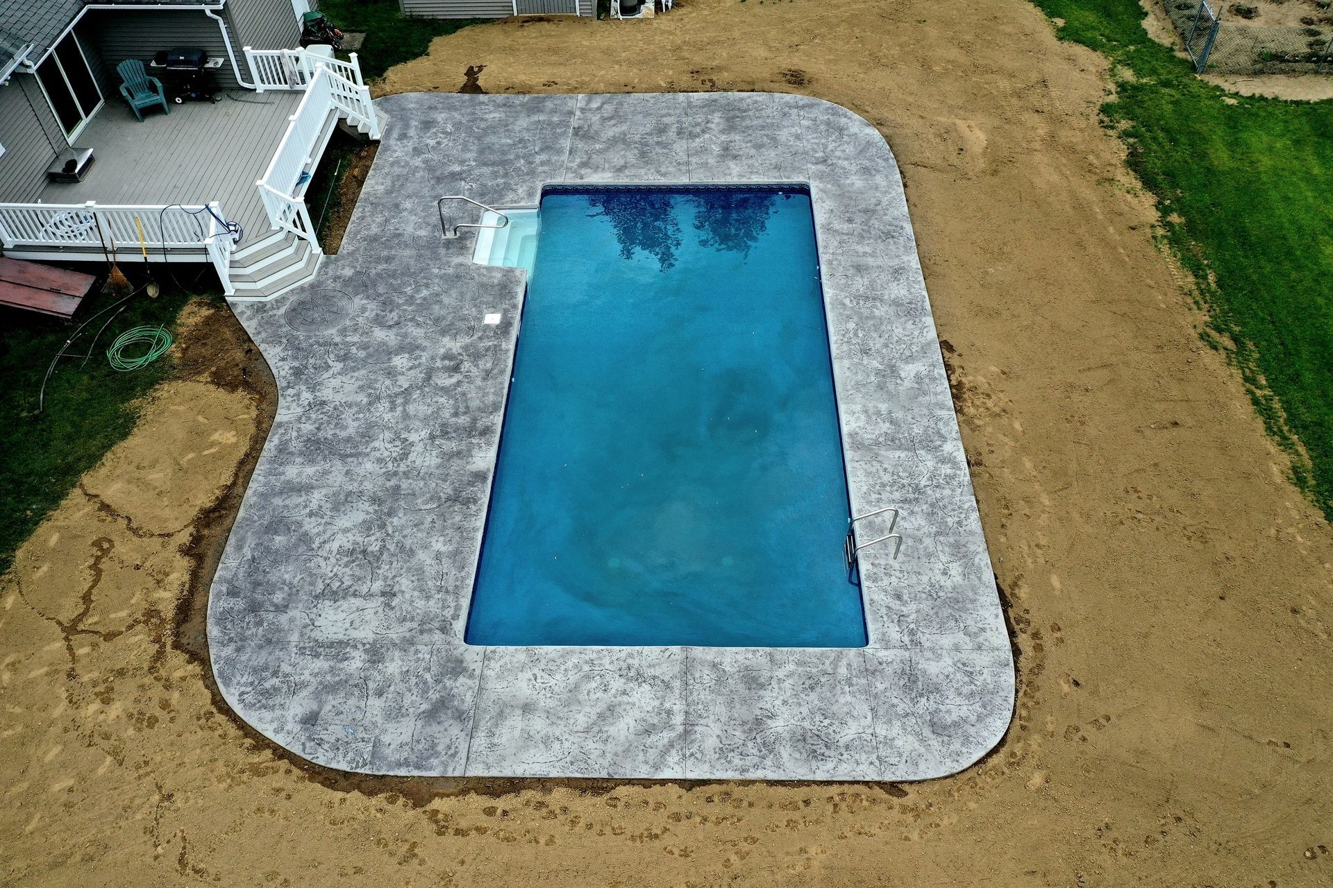 An aerial view of a rectangular blue swimming pool with a textured gray concrete deck, surrounded by unfinished dirt.