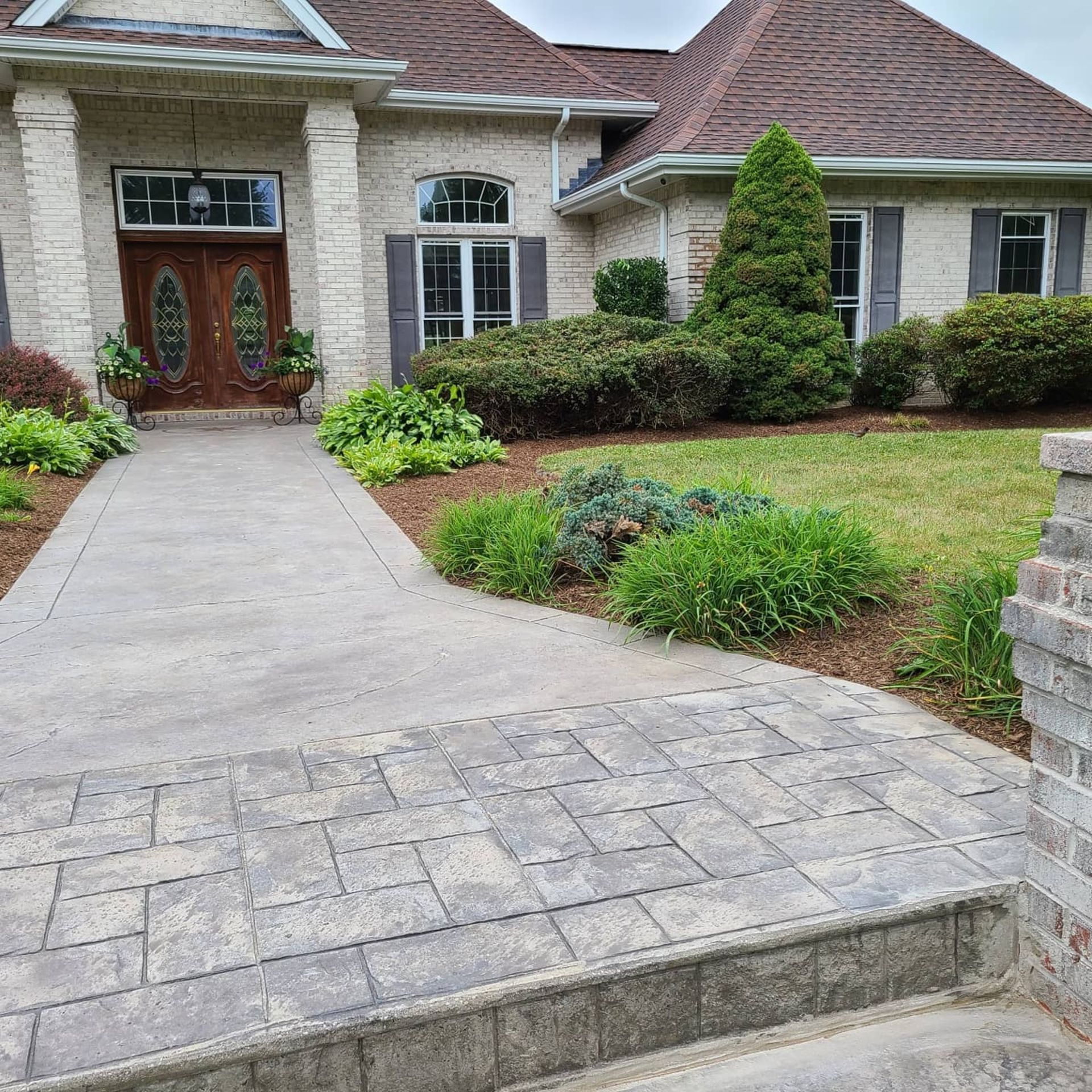 A beige stone house with a double wooden front door, a concrete walkway, and a landscaped front yard with green bushes.