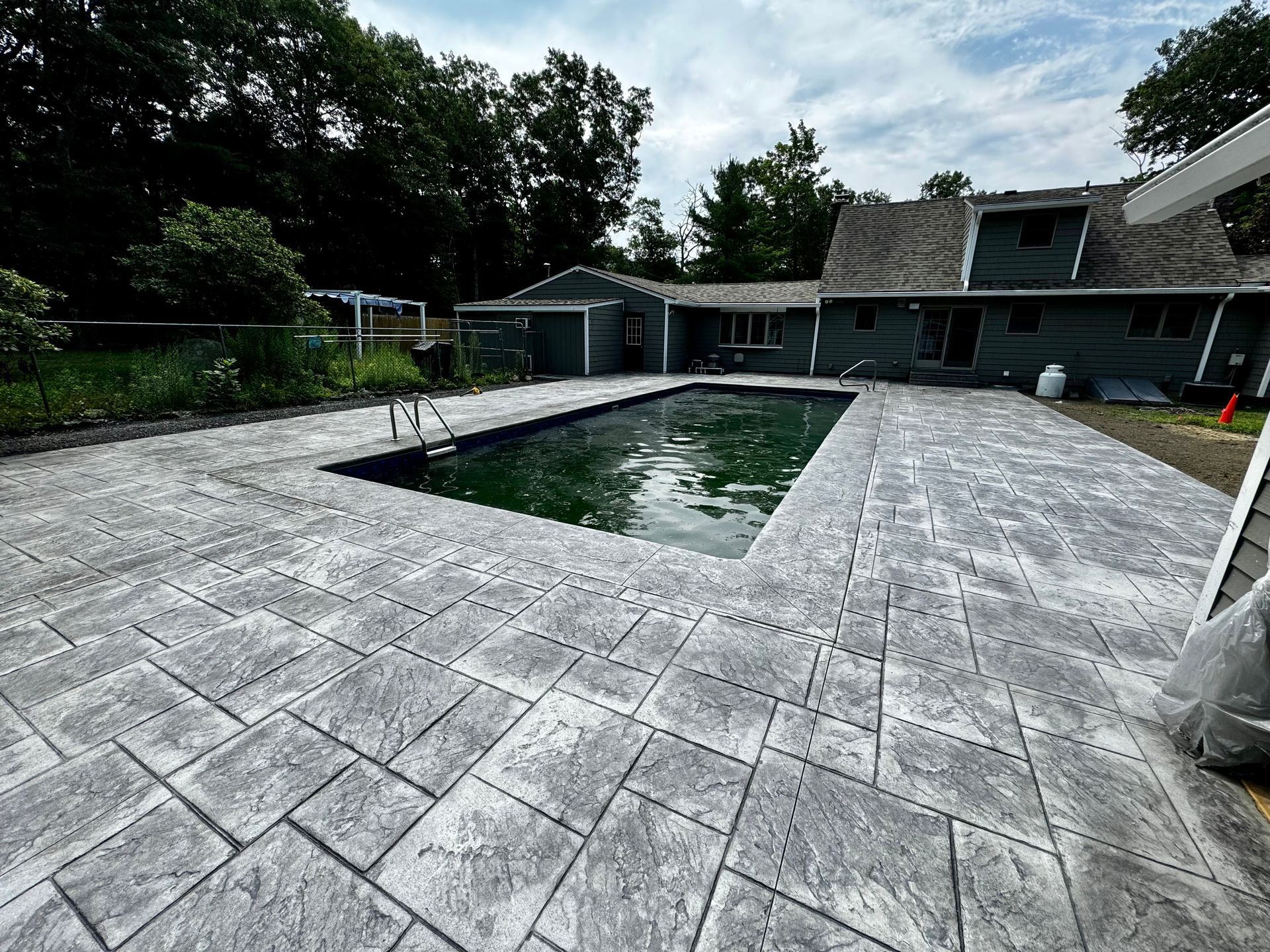 A rectangular swimming pool with green water surrounded by a gray, patterned concrete patio and a dark house in the back.