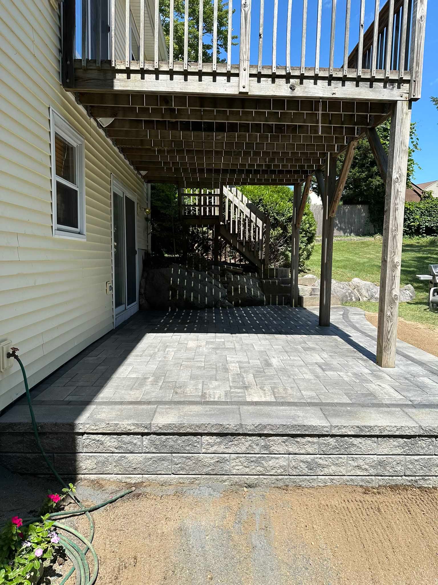 A newly installed gray paver patio beneath a wooden deck attached to the side of a light yellow house.