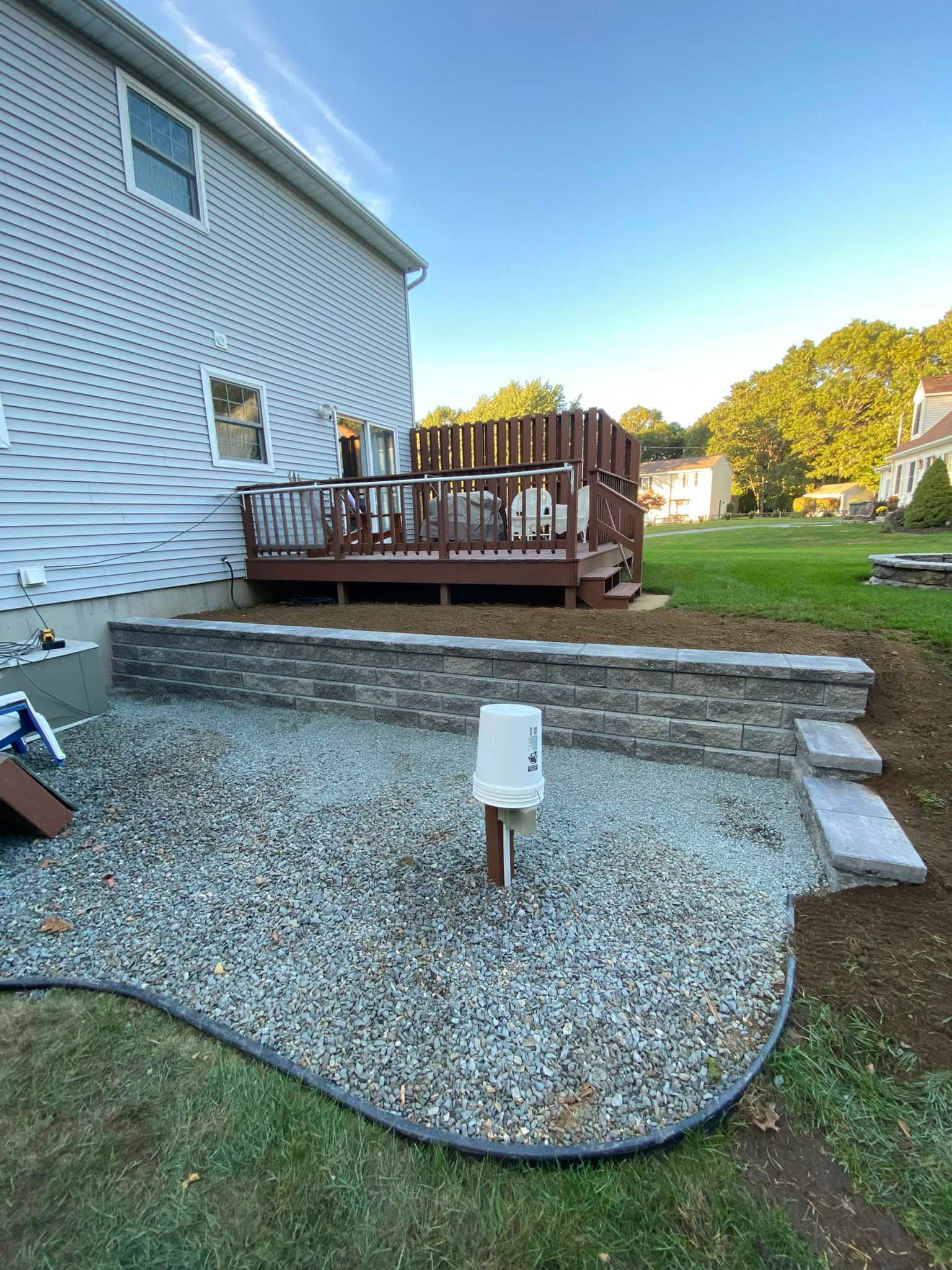 A gray house with a wooden deck overlooks a gravel patio area featuring a low, stone retaining wall and a white bucket.