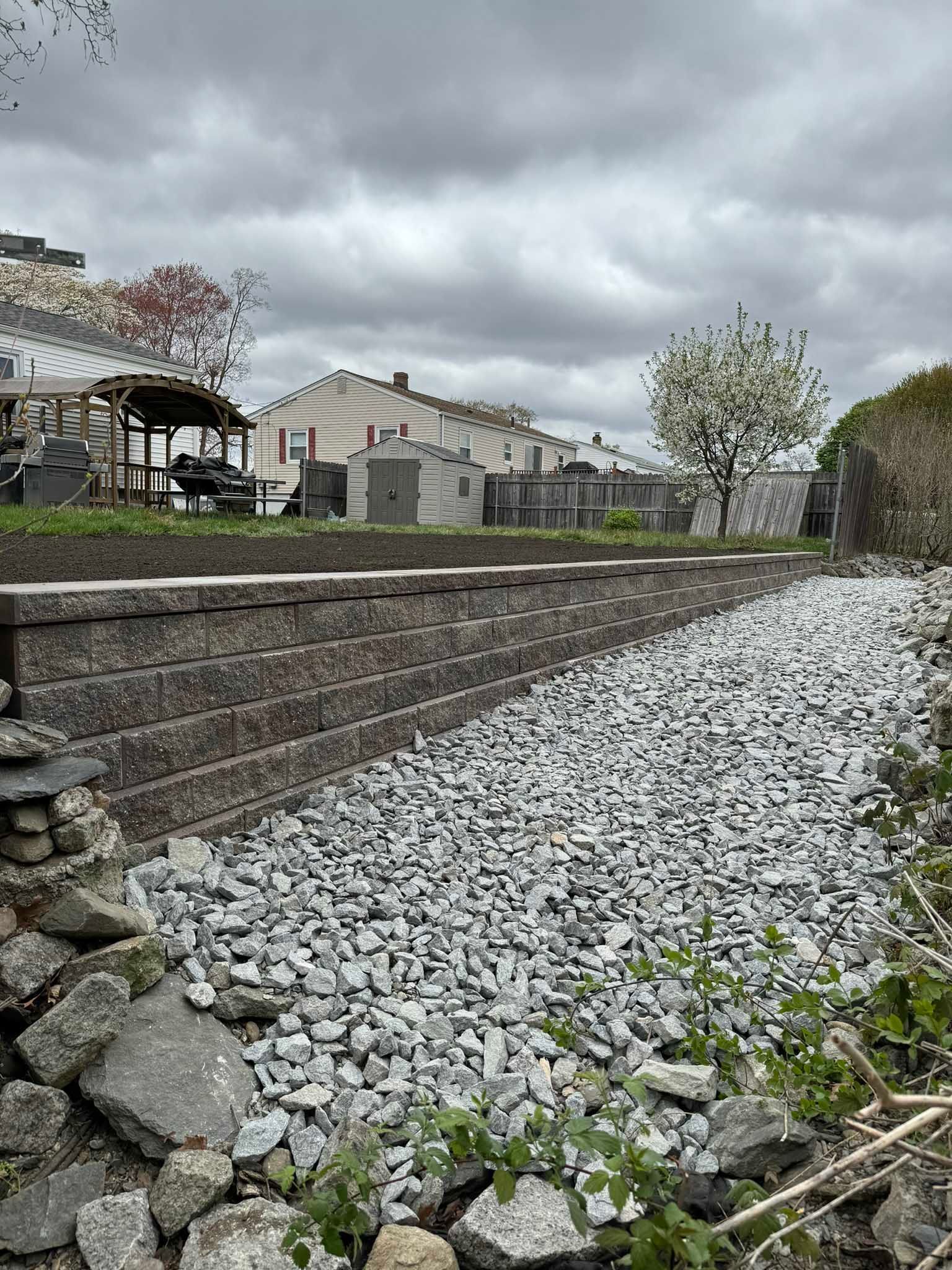 A landscaped backyard featuring a stone retaining wall bordered by a bed of light gray gravel under a cloudy sky.