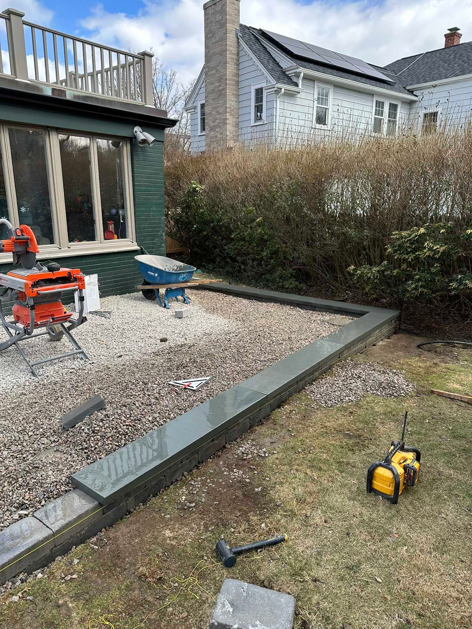 Construction site with gray stone edging, gravel patio, saw, and yard tools near a house with a deck.