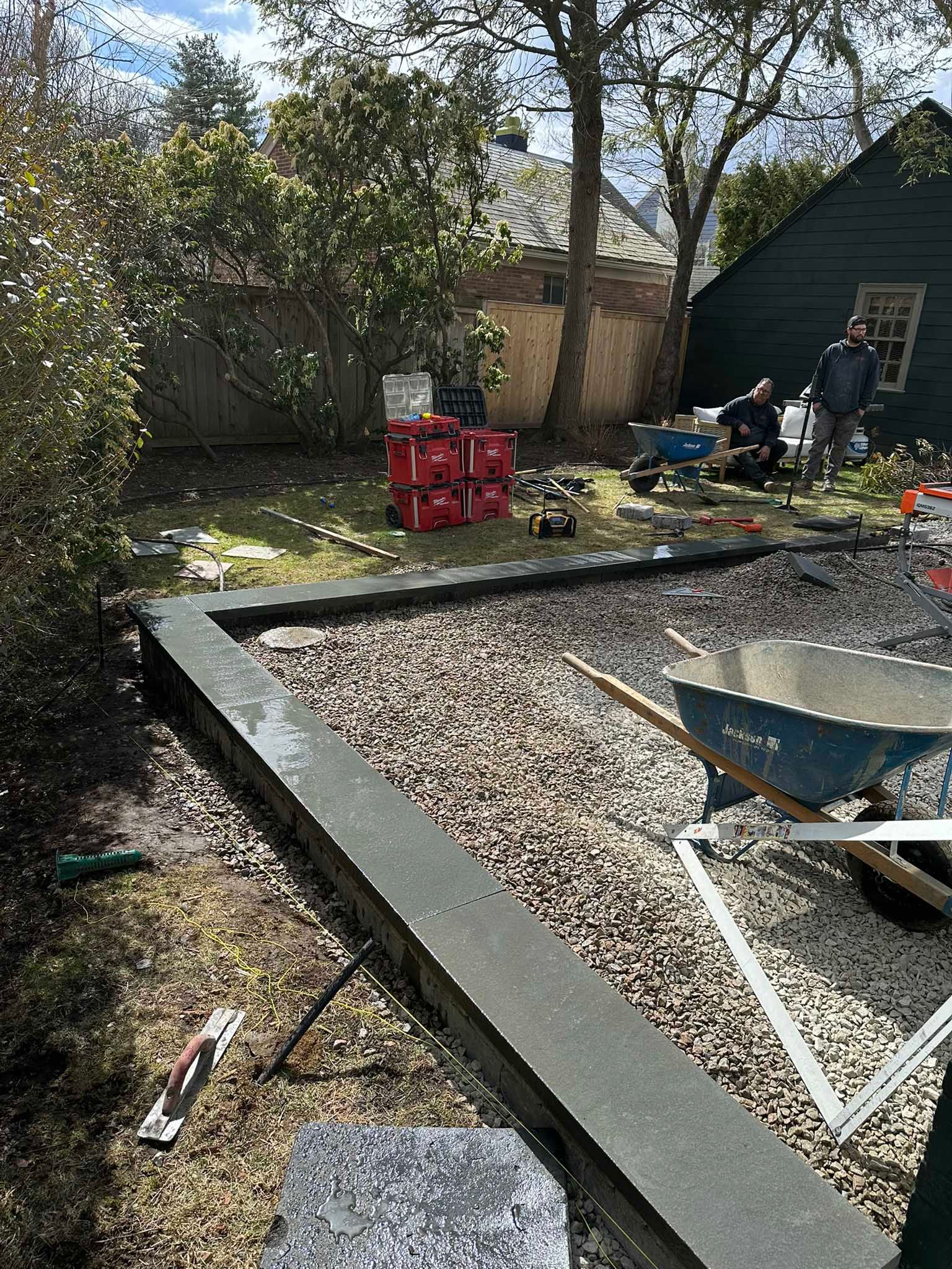 Workers build a stone-edged gravel patio in a residential backyard, with tools and red crates nearby.