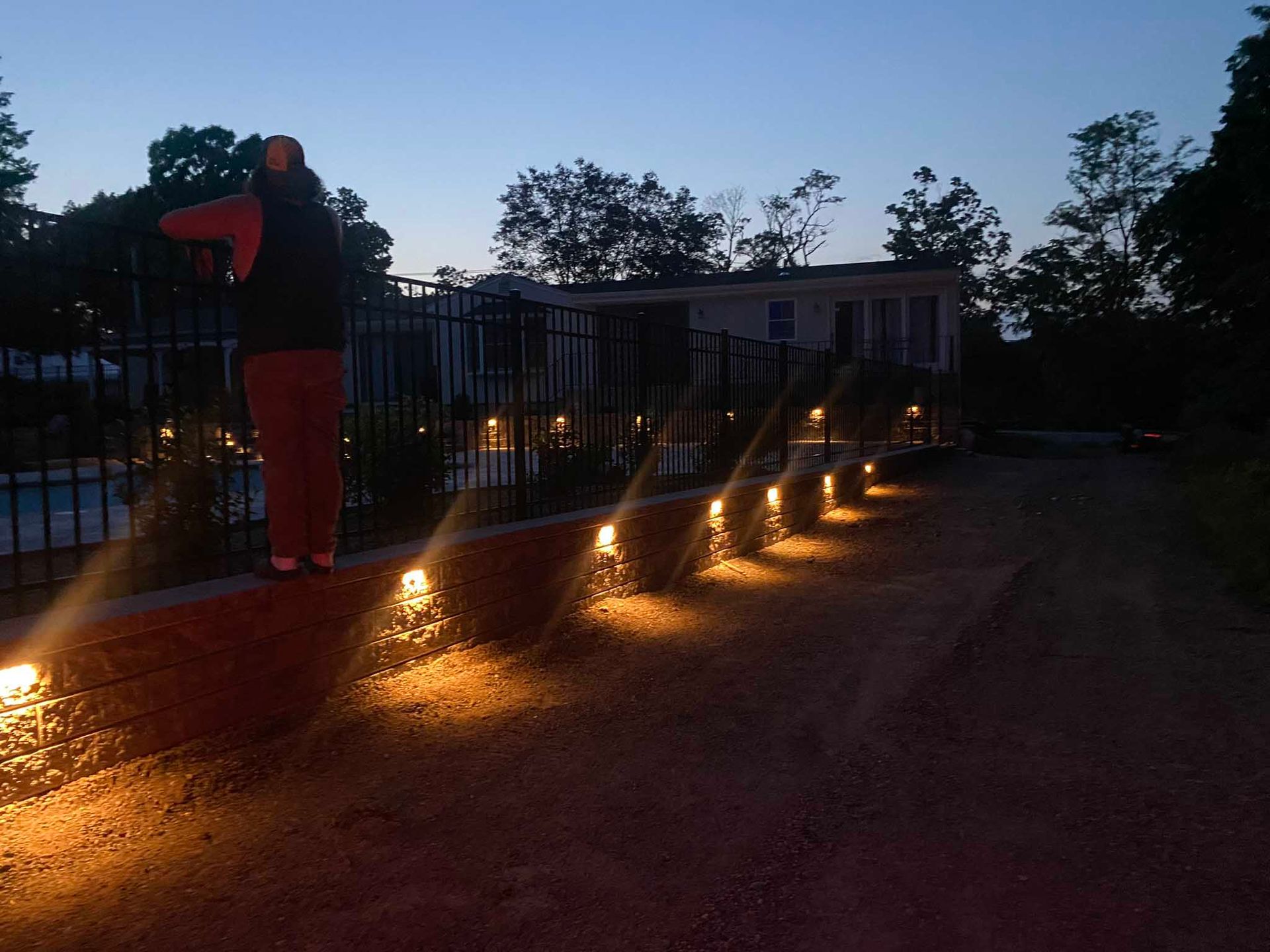 A person stands on a brick wall illuminated by a row of outdoor lights at twilight, next to a black fence and pool.