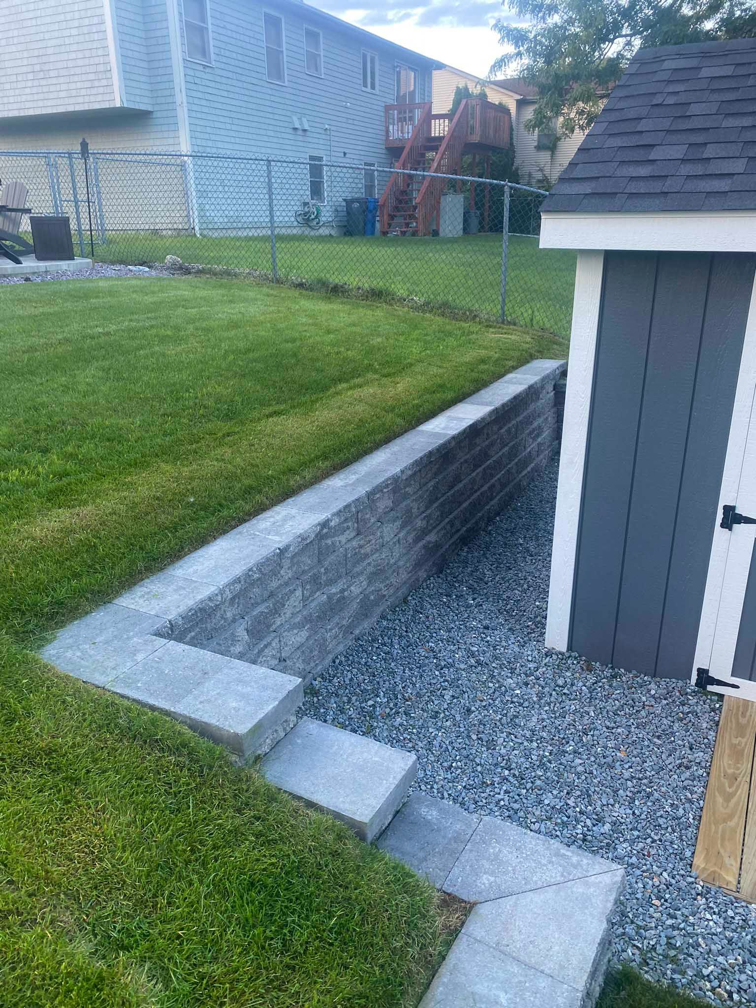 A stone retaining wall built into a grassy hill next to a gray storage shed, with a gravel-covered area in front.