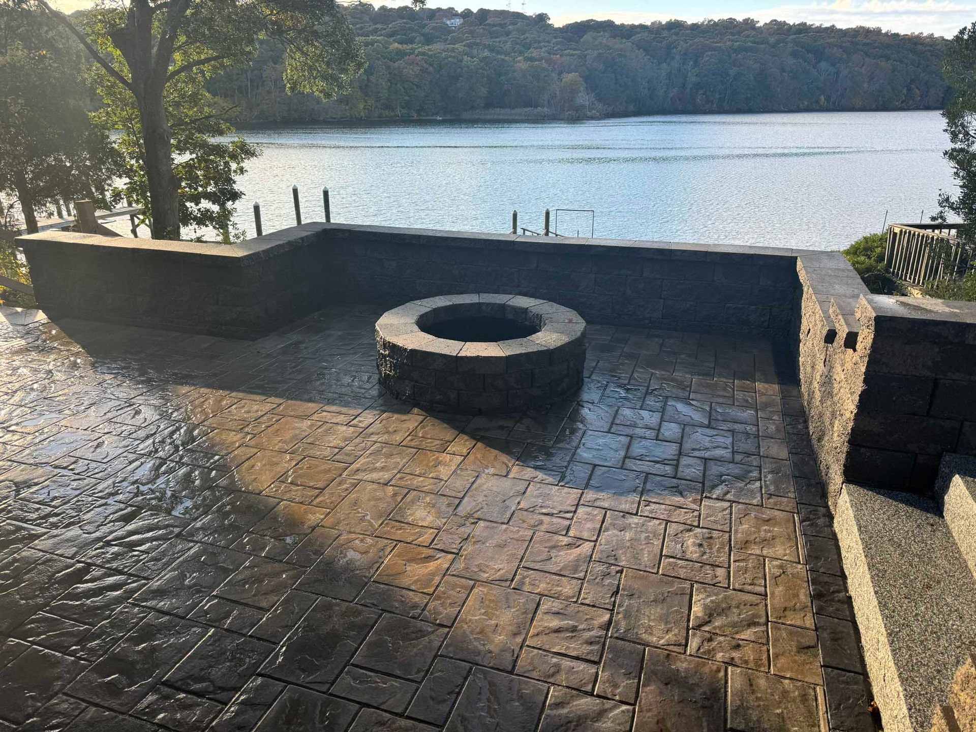 A stone patio with a central circular fire pit overlooking a lake surrounded by trees under a clear sky.