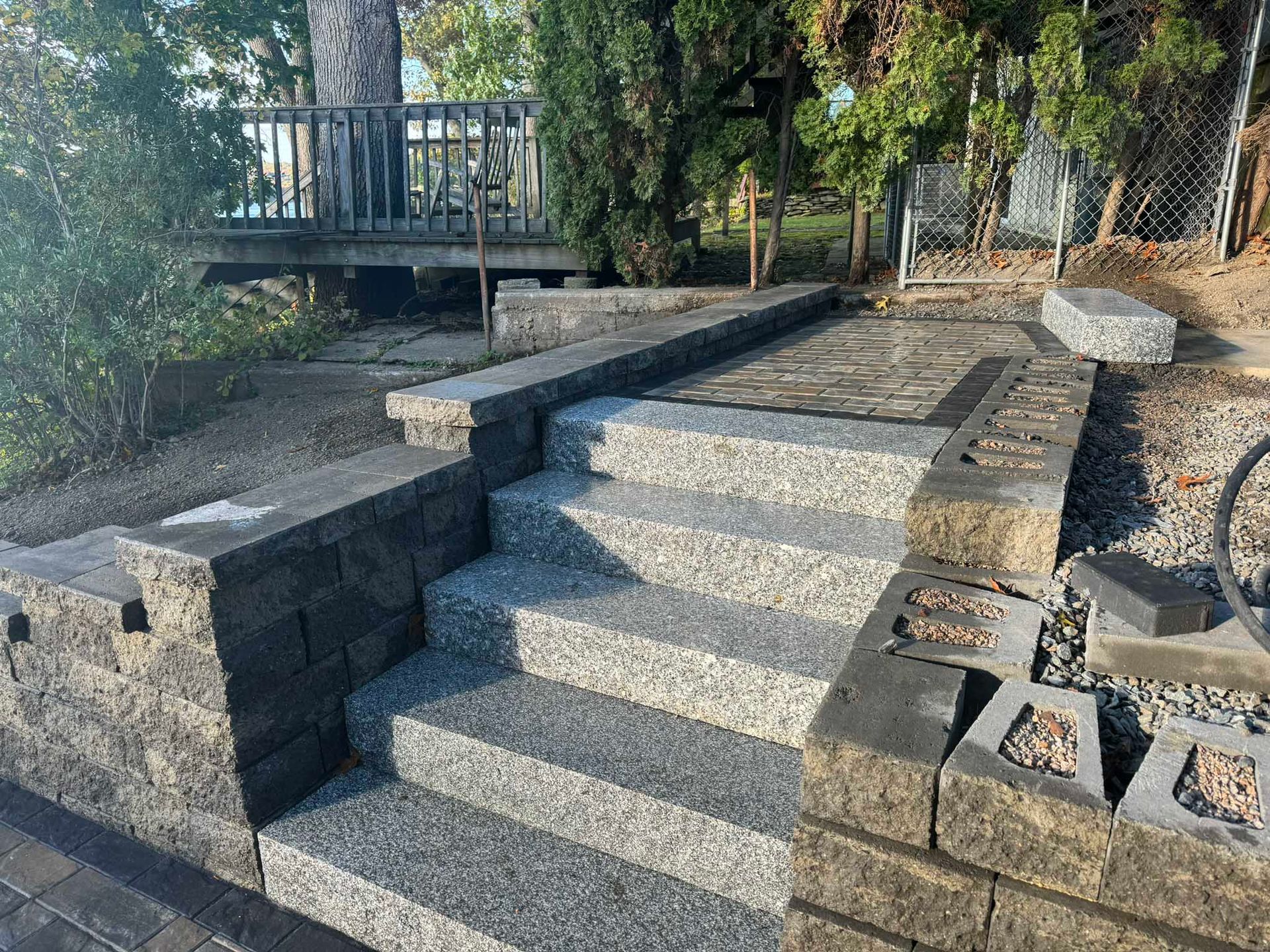 Stone steps with granite treads lead up to a tiered patio and wooden deck in a landscaped yard.