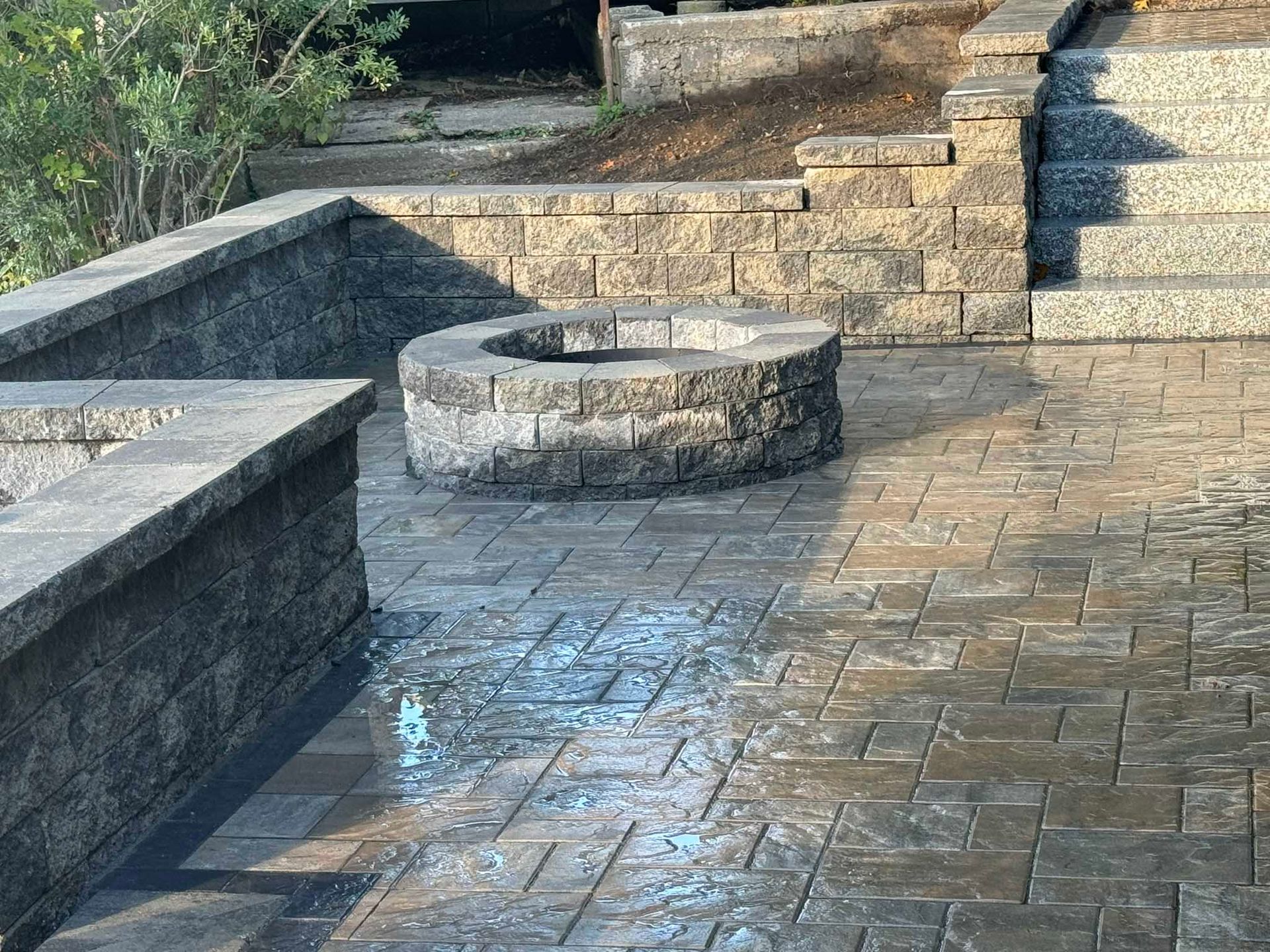 A circular stone fire pit sits in the center of a paved patio surrounded by low stone retaining walls and stairs.