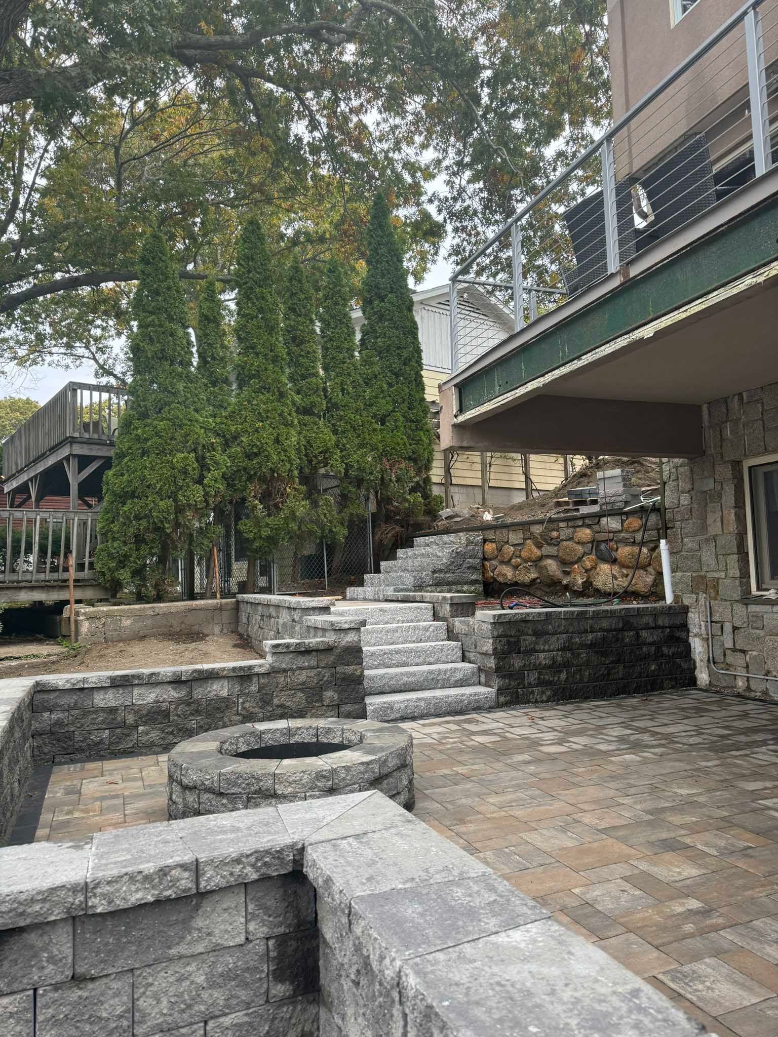 A gray paver patio with a stone fire pit, tiered stone retaining walls, and a stone staircase leading up a hillside.