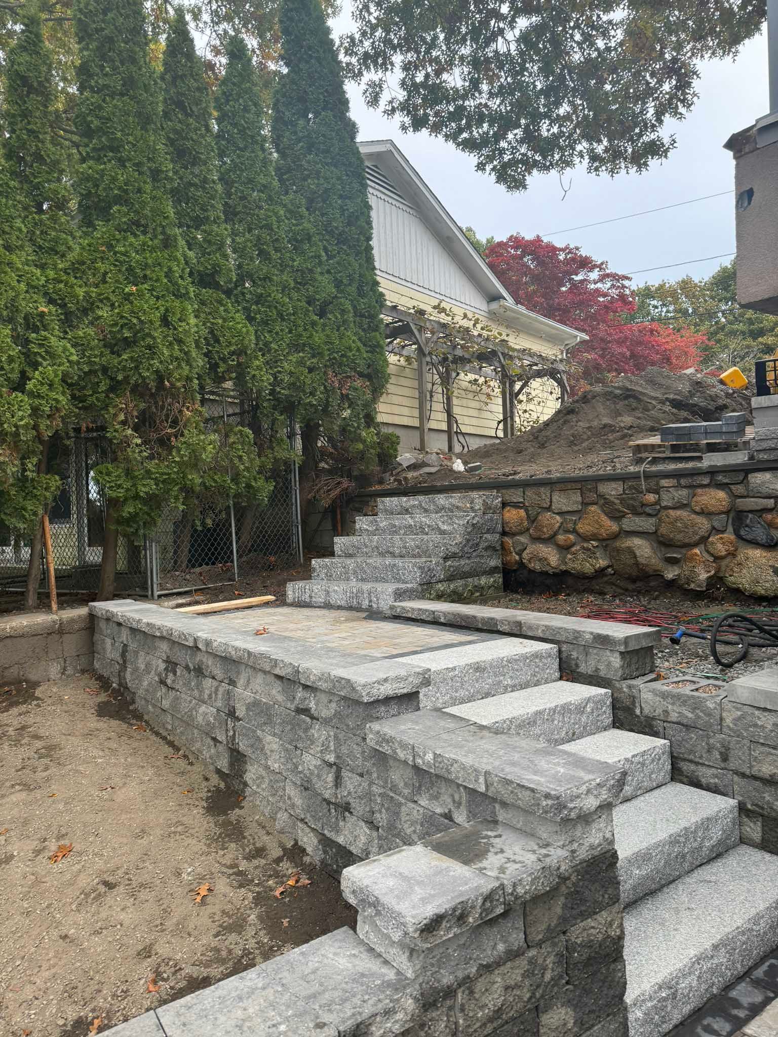 New gray stone tiered retaining walls and stairs under construction in a residential yard near mature evergreen trees.