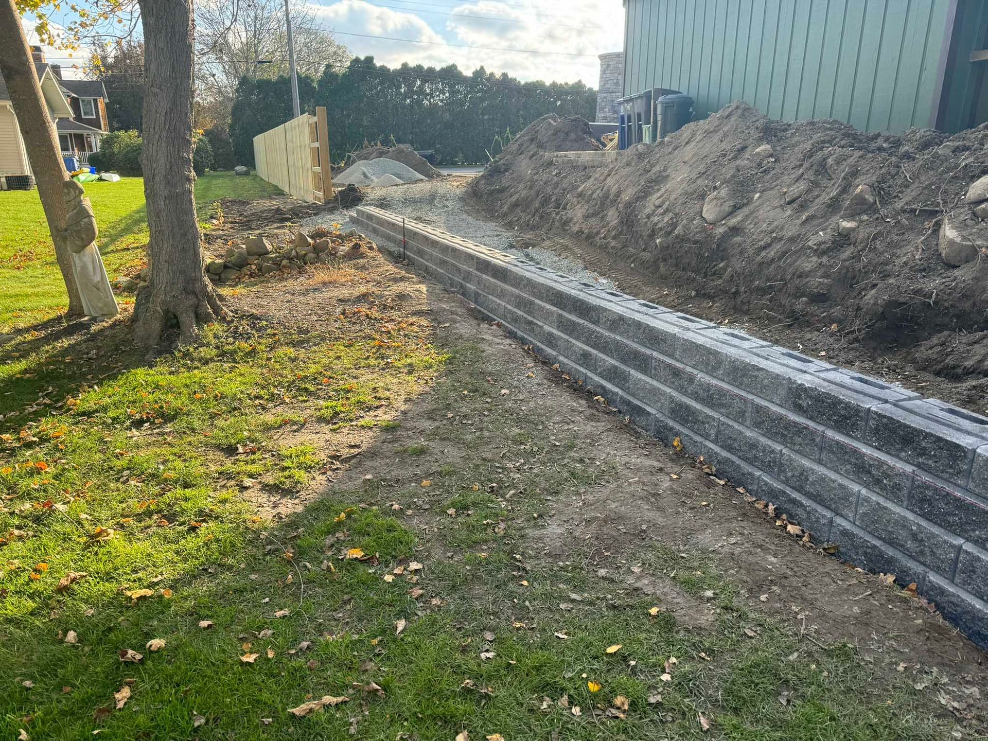 A partially constructed gray stone retaining wall separates a grassy yard from an excavated dirt mound near a building.
