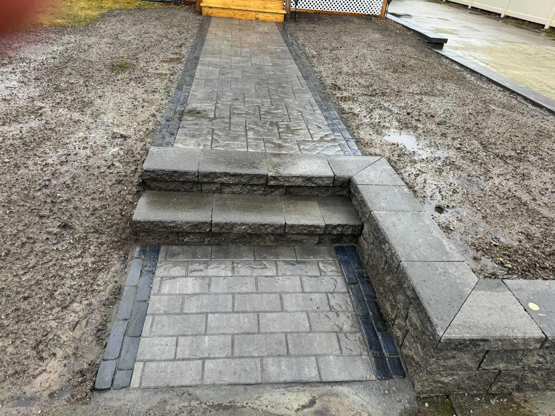 A gray paver walkway leads up two stone steps, bordered by a matching low stone retaining wall amidst muddy ground.