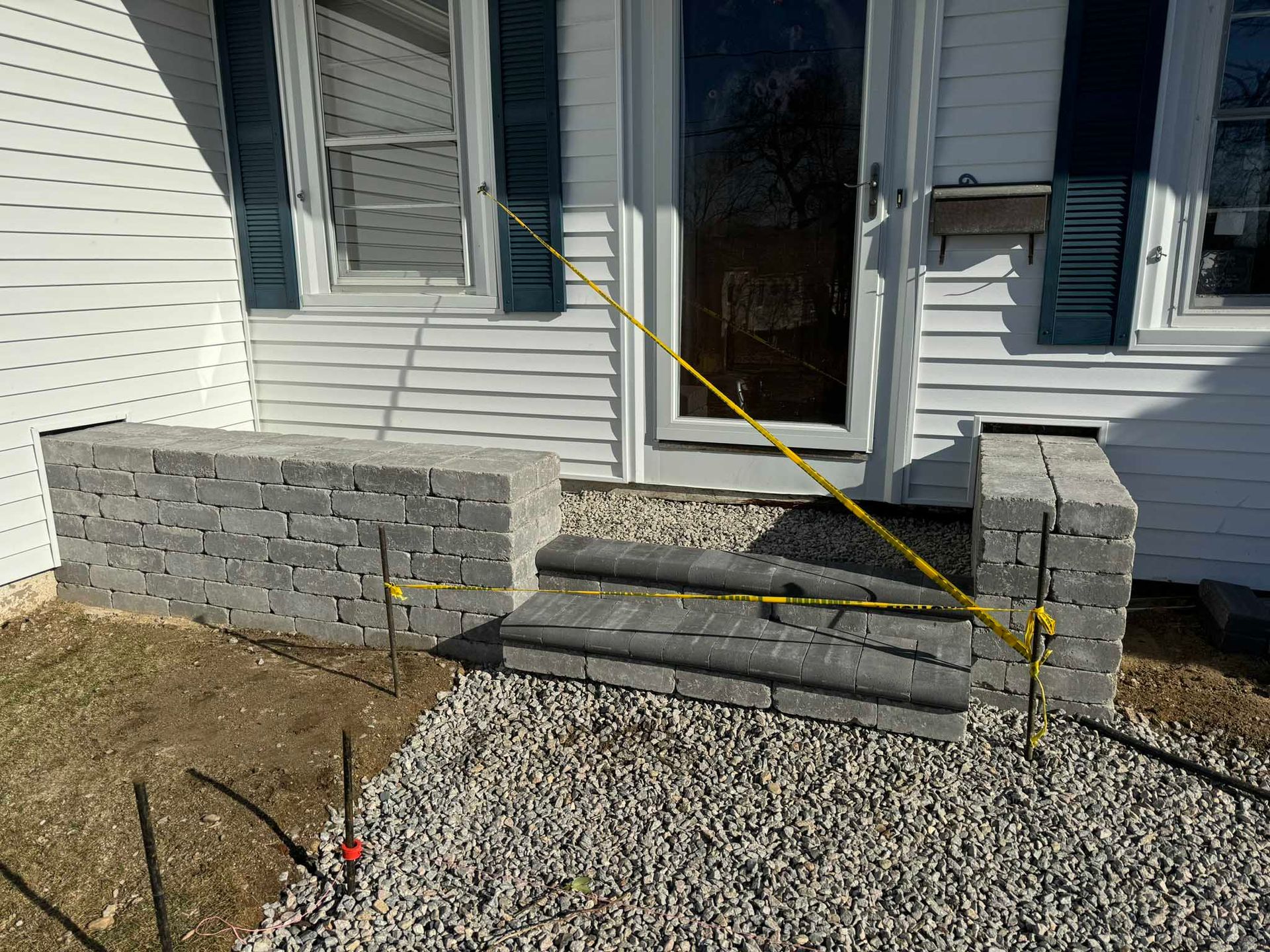 A partially constructed stone stoop with steps in front of a white house, marked off by yellow caution tape and stakes.