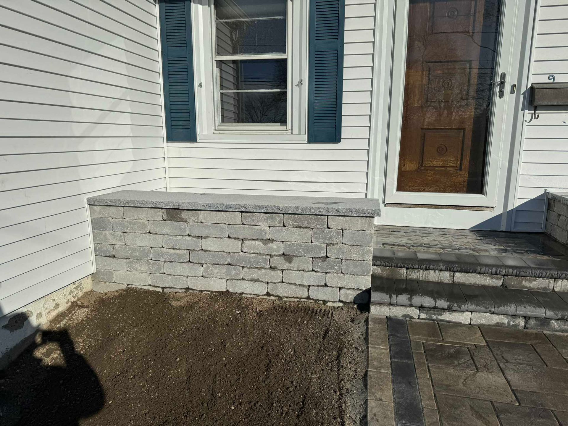 A low gray stone wall built against white siding next to a front door with a window and a paved walkway.