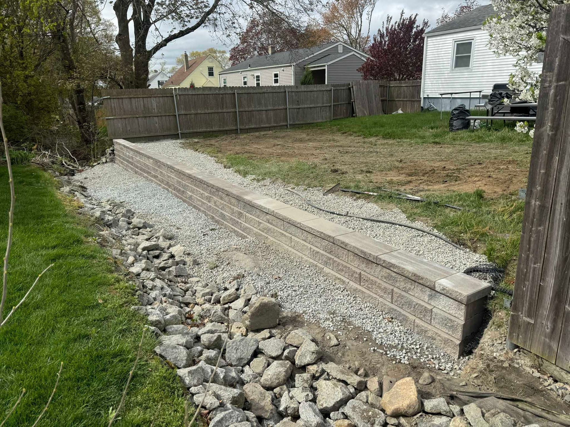 A newly constructed retaining wall made of light-colored stone blocks, bordered by gravel and a rocky drainage area.