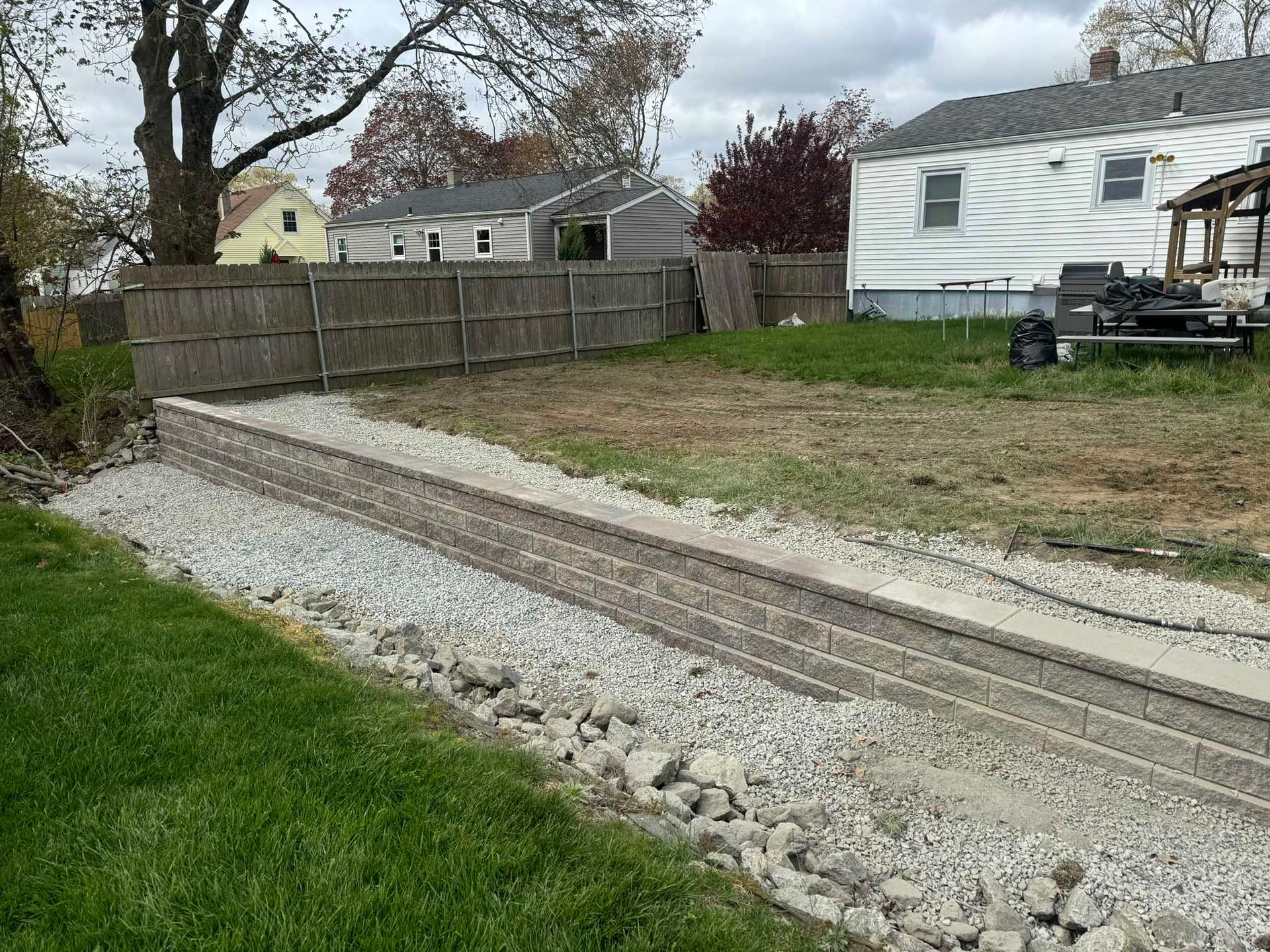 A backyard landscape featuring a newly built gray stone retaining wall separating a gravel path from a grassy upper yard.